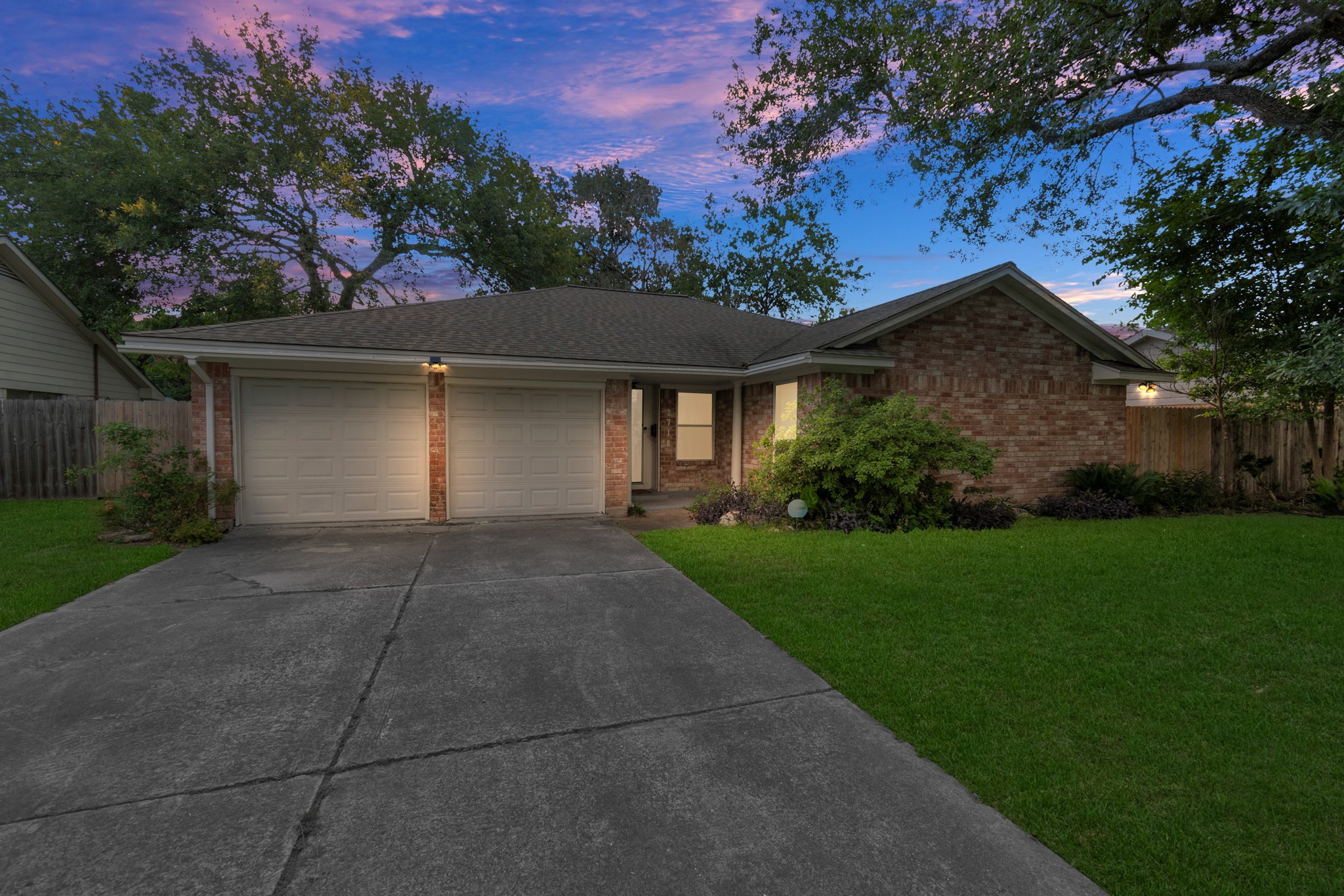 Charming brick home with great curb appeal. Features a well-kept front yard, mature trees, and a wide driveway leading to a two-car garage. Warm exterior lighting adds a welcoming feel in the evening.