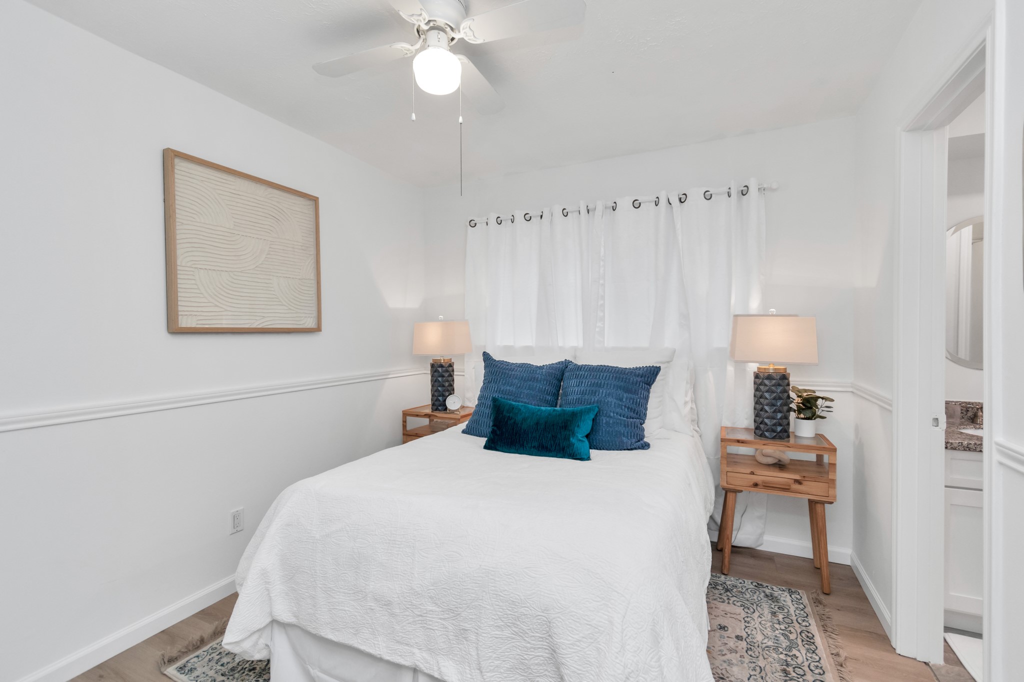2718 Lazy Spring Drive Houston, TX 77080 - Photo 20 of 32 This cozy bedroom features a comfortable bed with crisp white linens, accented by blue throw pillows. It's illuminated by stylish lamps on wooden nightstands, with a ceiling fan overhead. The room has a modern, minimalist feel with a simple wall art piece and light wood flooring.