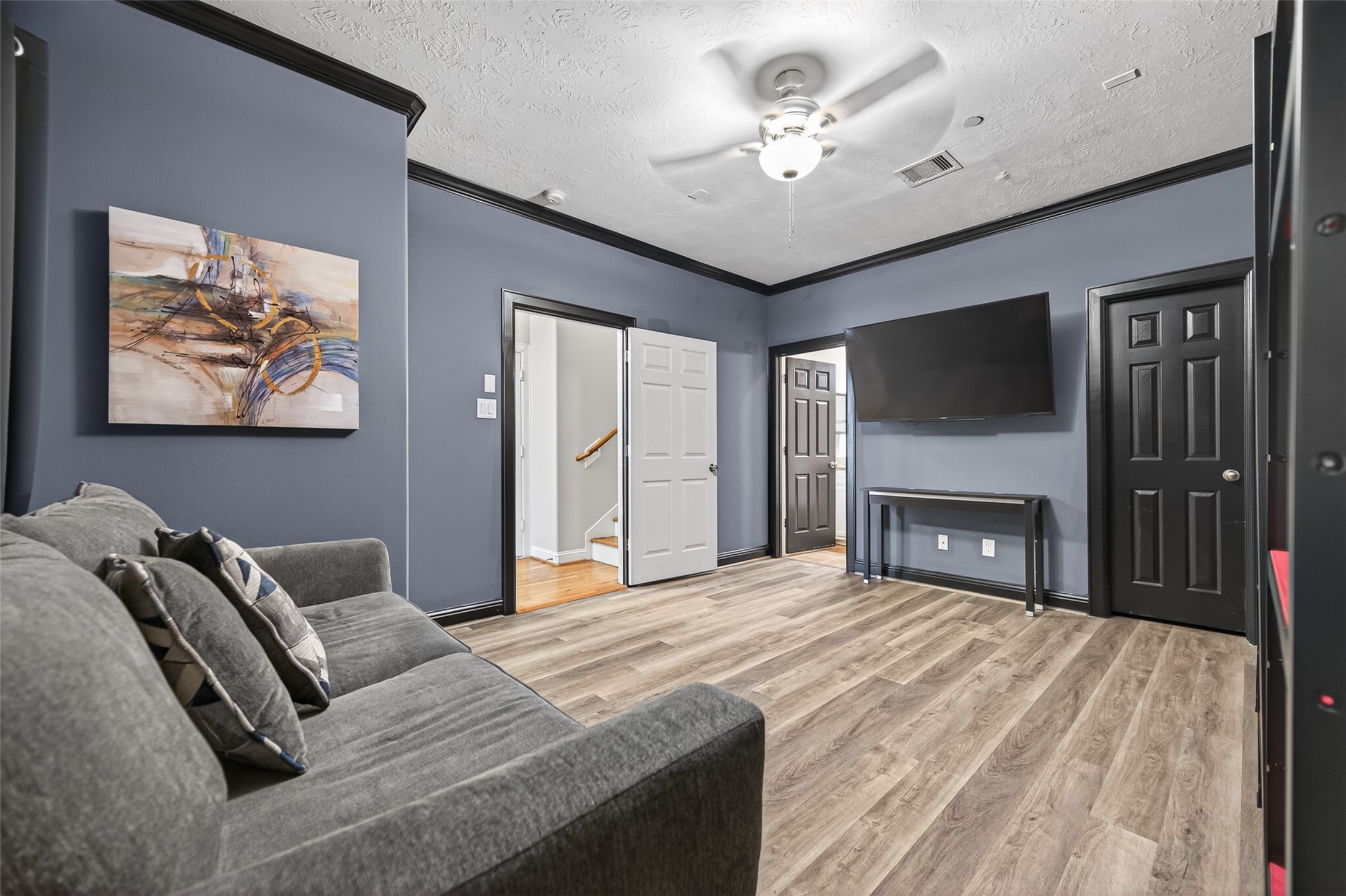 2611 Riverside Drive, Unit C Houston, TX 77004 - Photo 41 of 50 This stylish first-floor room showcases modern design with rich blue-gray walls, bold black trim, and wood-look flooring. A sleek ceiling fan, gray sofa, wall-mounted TV, and abstract artwork create a cozy yet contemporary atmosphere. Complete with a private en suite and a large closet, this versatile space was previously used as a photography and film studio, offering endless possibilities for use.