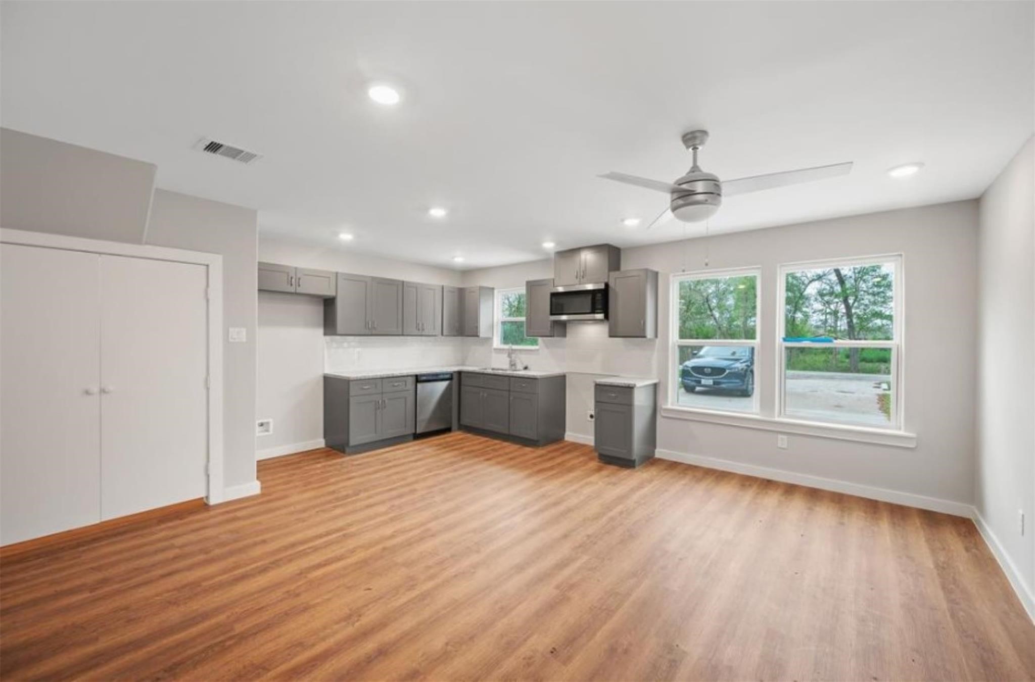 8120 Laura Koppe Road, Unit B Houston, TX 77028 - Photo 4 of 25 This photo showcases a modern open-plan kitchen and living area with wood flooring, gray cabinetry, and ample natural light from large windows. The space features recessed lighting and a ceiling fan, creating a bright and airy atmosphere.