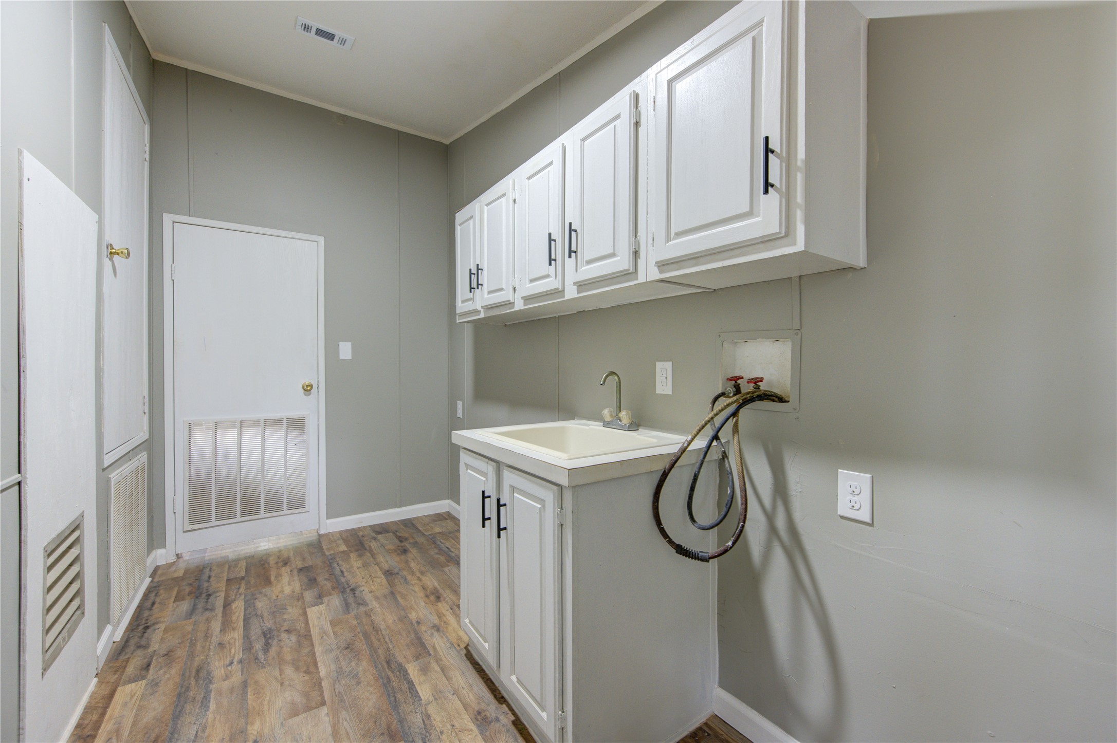 30226 Davis Street Magnolia, TX 77355 - Photo 27 of 50 This photo shows a laundry room with light-colored walls and wood-style flooring. It features white cabinets for storage, a utility sink with hook-ups for a washer, and a door leading to another area.