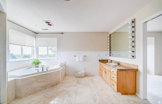1273 Grayson Court Sandy, UT 84094 - Photo 17 of 27 Spacious bathroom featuring a corner soaking tub with surrounding windows, dual vanity with a stone countertop, large mirror with vanity lighting, and light-toned tile flooring