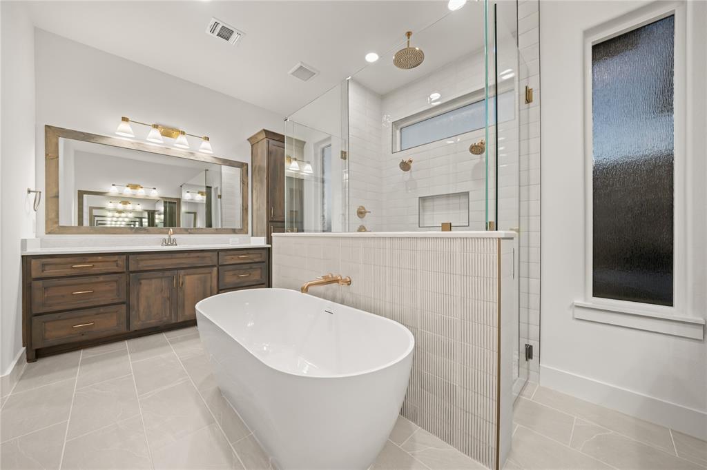1031 Ranch Vista Road Granbury, TX 76049 - Photo 24 of 32 Freestanding soaking tub with gold-finish fixtures, a spacious walk-in shower with dual shower heads, a long vanity with wood-finish cabinetry, a large framed mirror, and light tile flooring