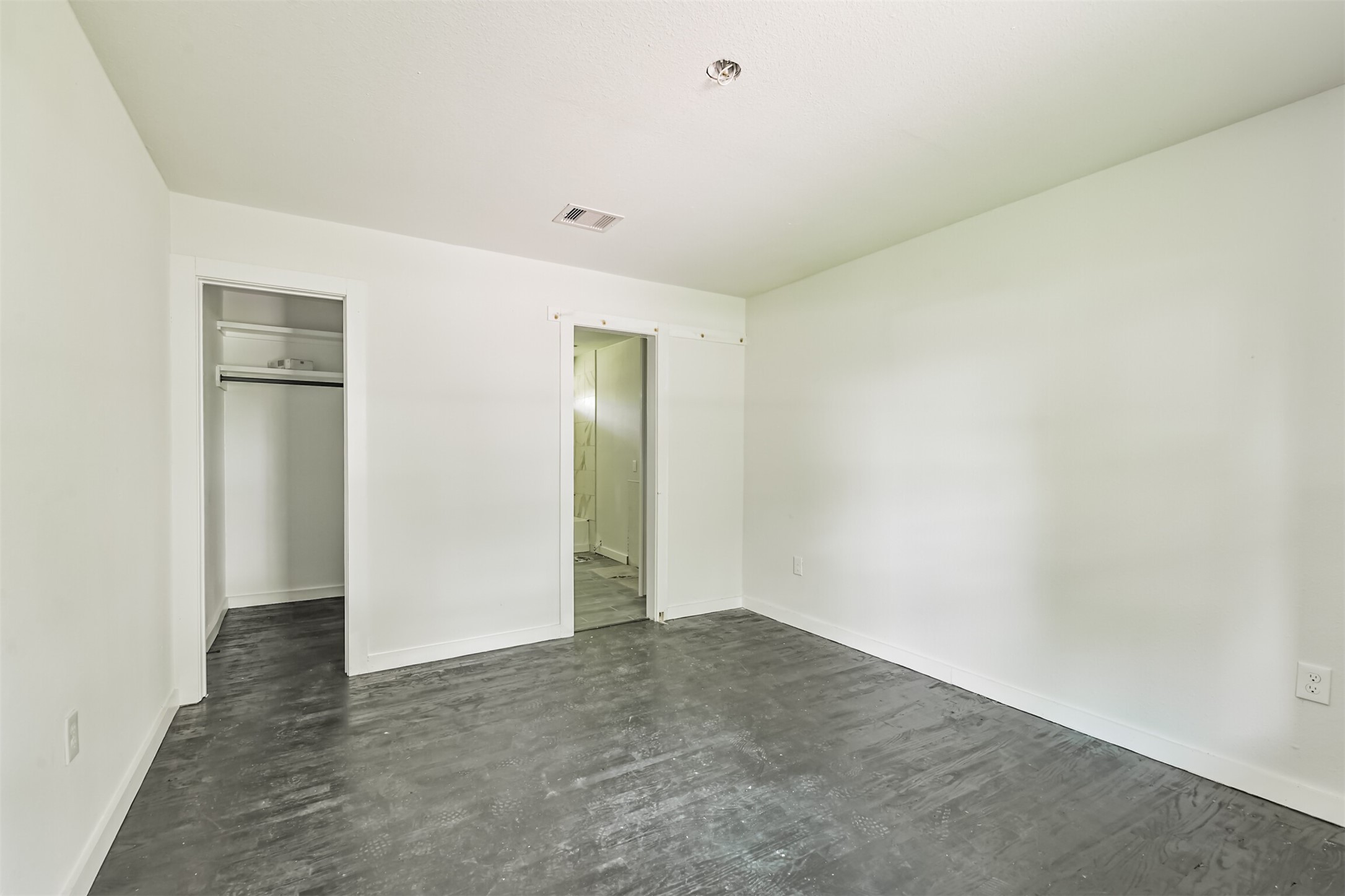 209 Leago Street Houston, TX 77022 - Photo 11 of 24 This photo shows a bright, minimalist room with light-colored walls and dark flooring. It features an open closet space and an adjoining door leading to a bathroom, creating a functional layout.