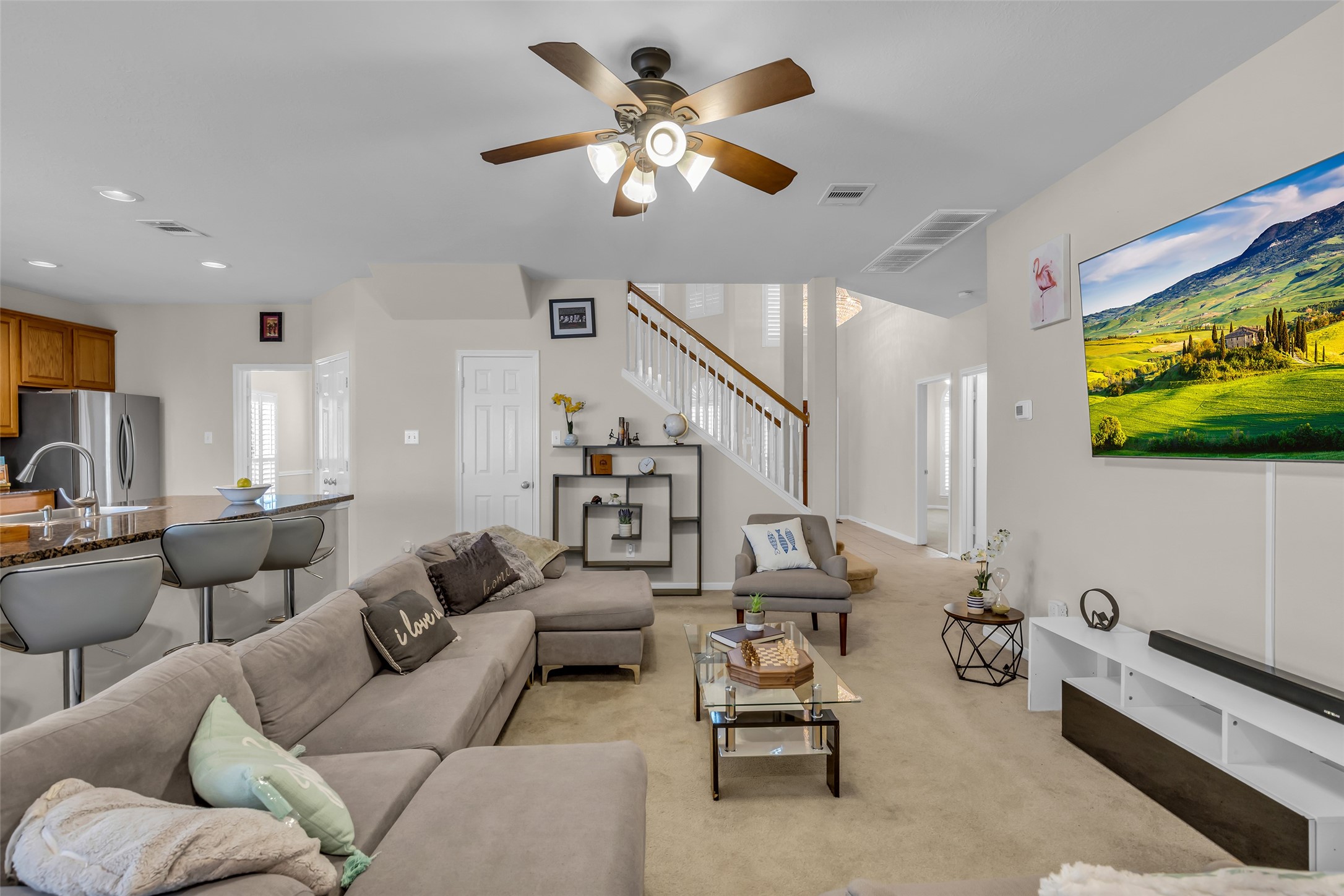 6807 Chapelfield Lane Houston, TX 77049 - Photo 24 of 42 This photo showcases a spacious, open-plan living area with neutral tones, featuring a comfortable sectional sofa and a wall-mounted TV. The room is well-lit with a ceiling fan and recessed lighting. There's a modern kitchen in the background with a breakfast bar and stainless steel appliances. A staircase leads to the upper level, adding to the home's charm.