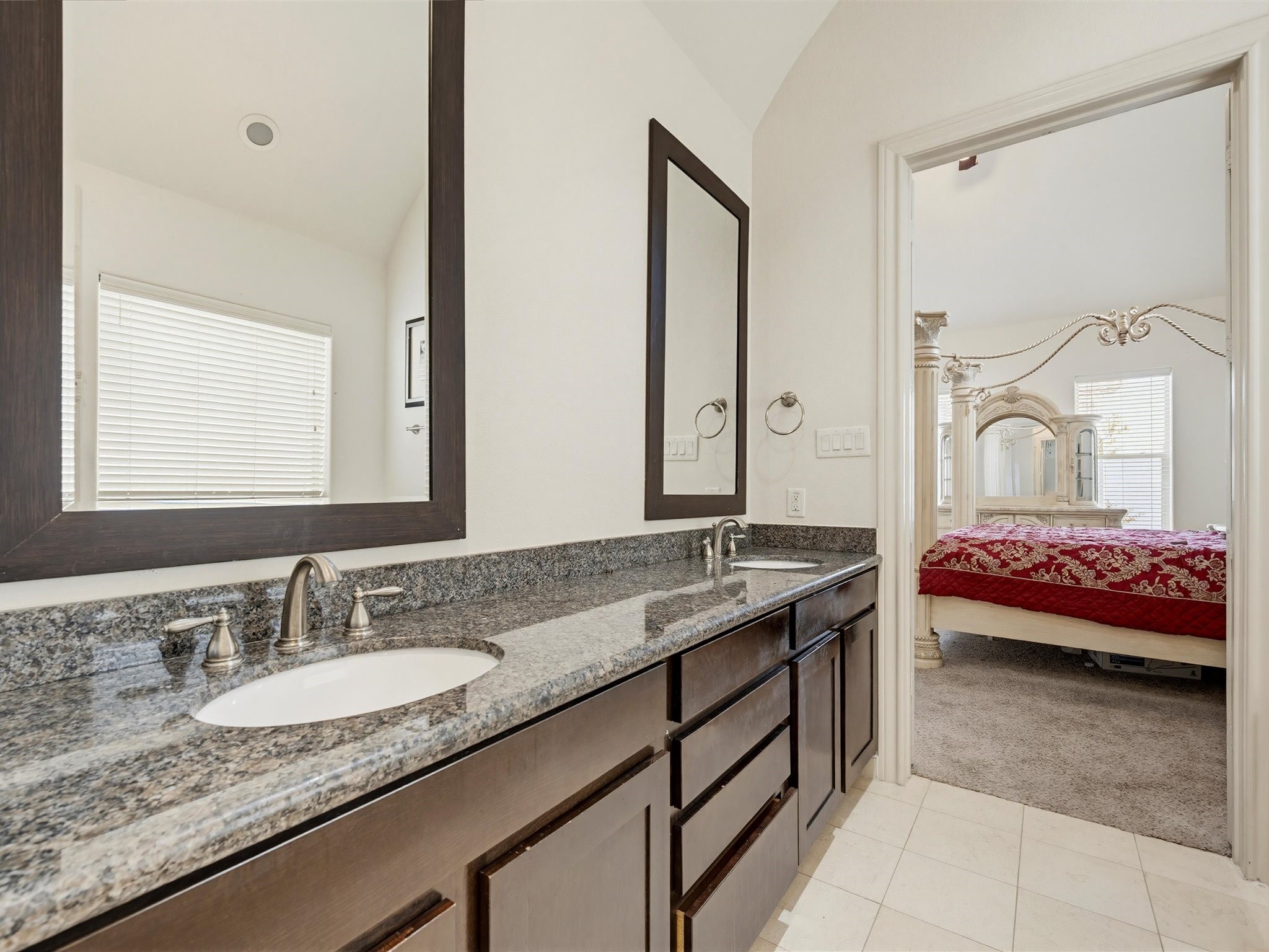 9427 London Bridge Station Houston, TX 77045 - Photo 24 of 30 This photo features a modern bathroom with dual sinks, granite countertops, and dark wood cabinetry. It connects to a bedroom with plush carpet and classic furniture, creating a seamless flow between spaces.