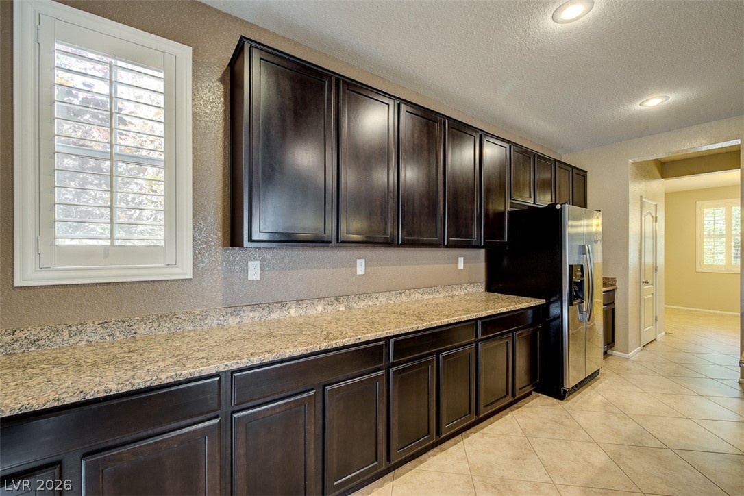 3199 Subtle Color Avenue Henderson, NV 89044 - Photo 15 of 46 Kitchen with dark wood finish cabinetry, light stone countertops, stainless steel fridge with ice dispenser, light tile patterned floors, and a textured ceiling