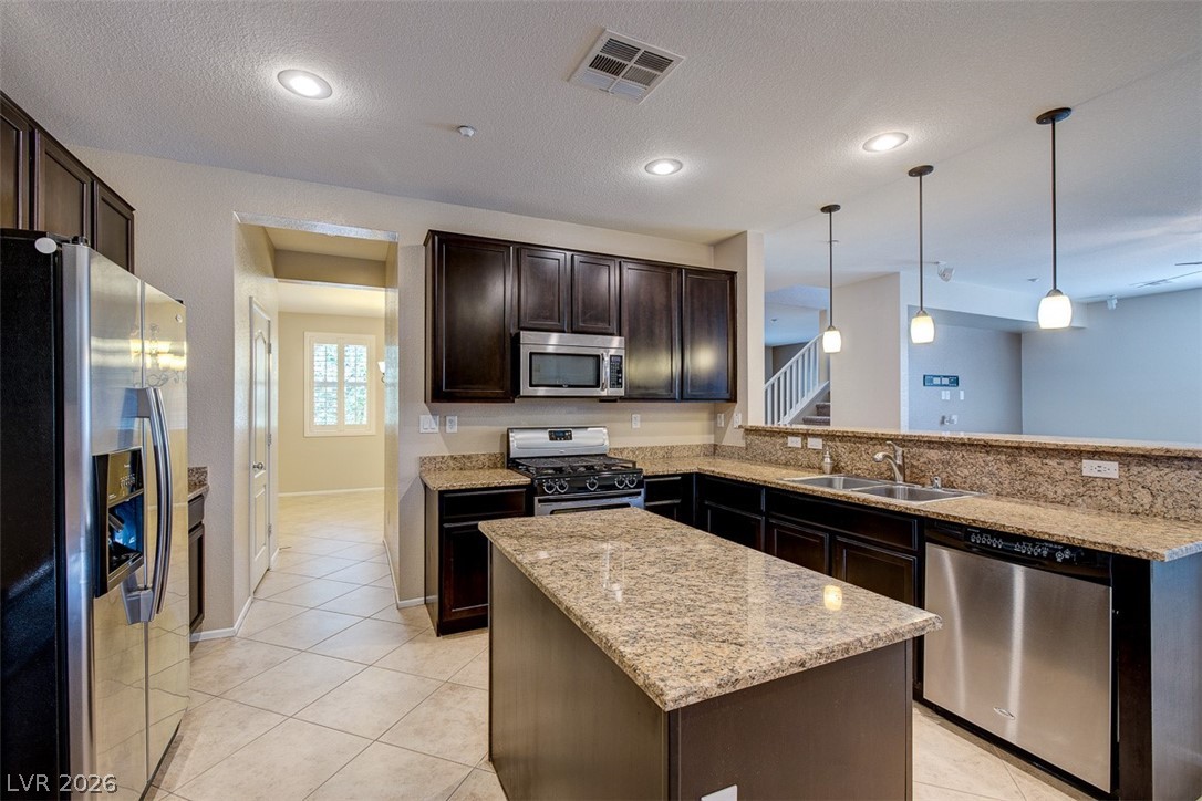 3199 Subtle Color Avenue Henderson, NV 89044 - Photo 16 of 46 Kitchen with a peninsula, stainless steel appliances, dark wood finish cabinetry, light stone countertops, and a center island