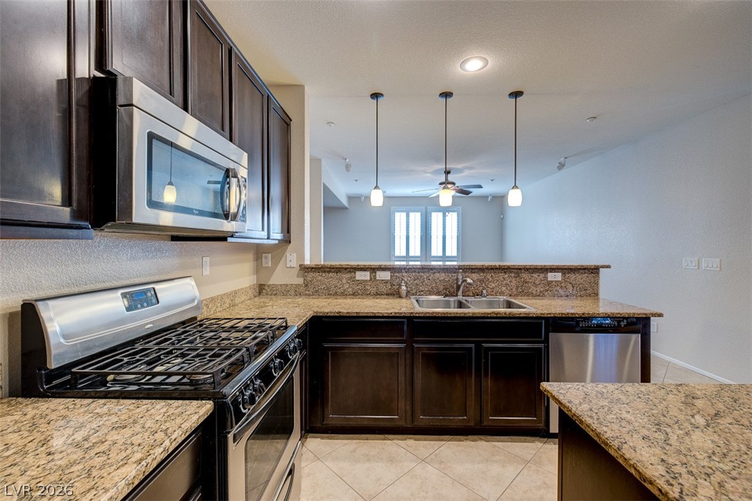 3199 Subtle Color Avenue Henderson, NV 89044 - Photo 17 of 46 Kitchen featuring stainless steel appliances, dark wood finish cabinetry, light stone counters, light tile patterned floors, and hanging light fixtures