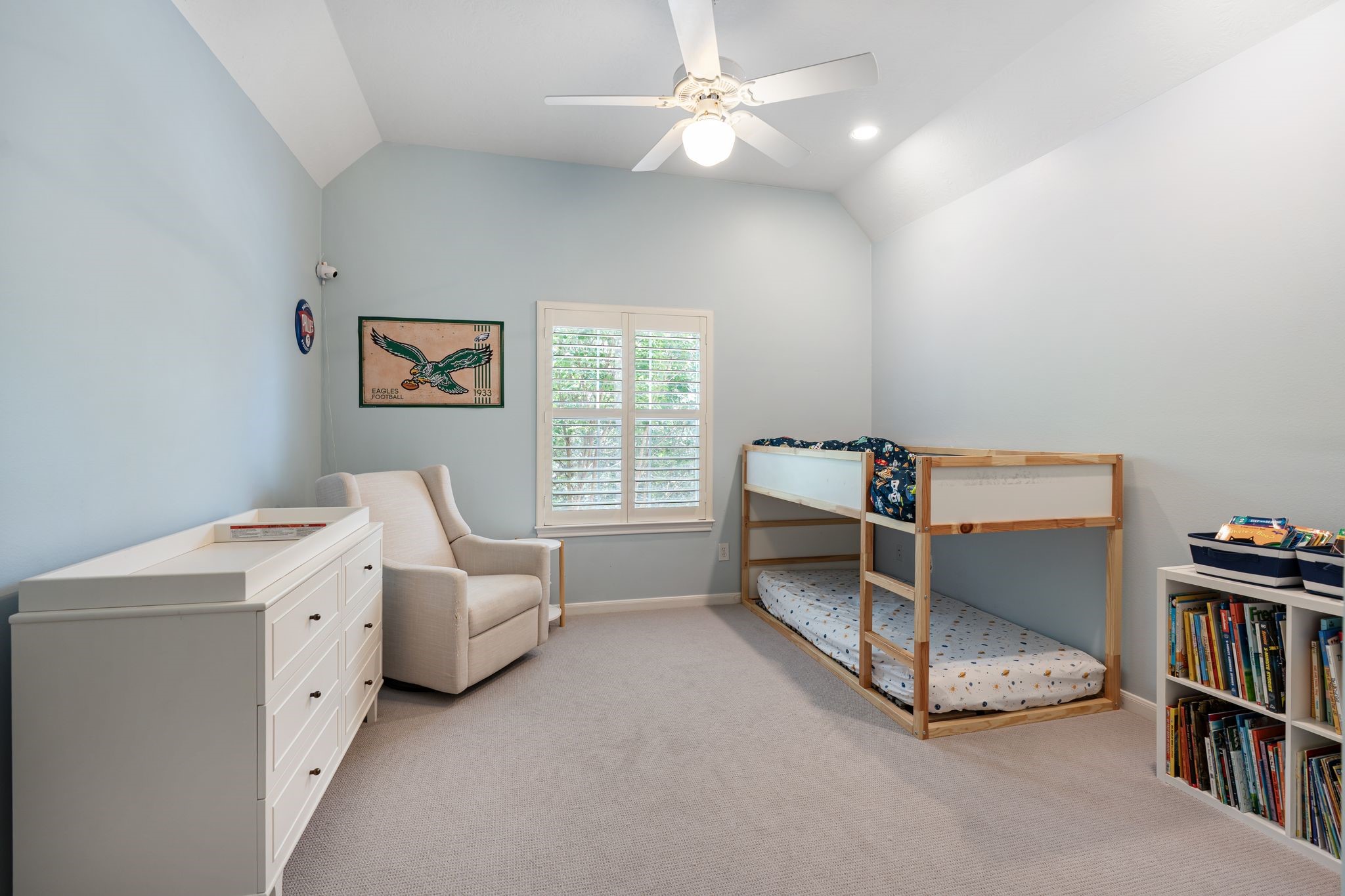 3822 Merrick Street Houston, TX 77025 - Photo 27 of 34 Another upstairs bedroom features a vaulted ceiling, carpet flooring, a ceiling fan, and a plantation shutter window with a view to the greenery outside. The room's soft blue walls and well-proportioned layout make it a comfortable and welcoming secondary suite.