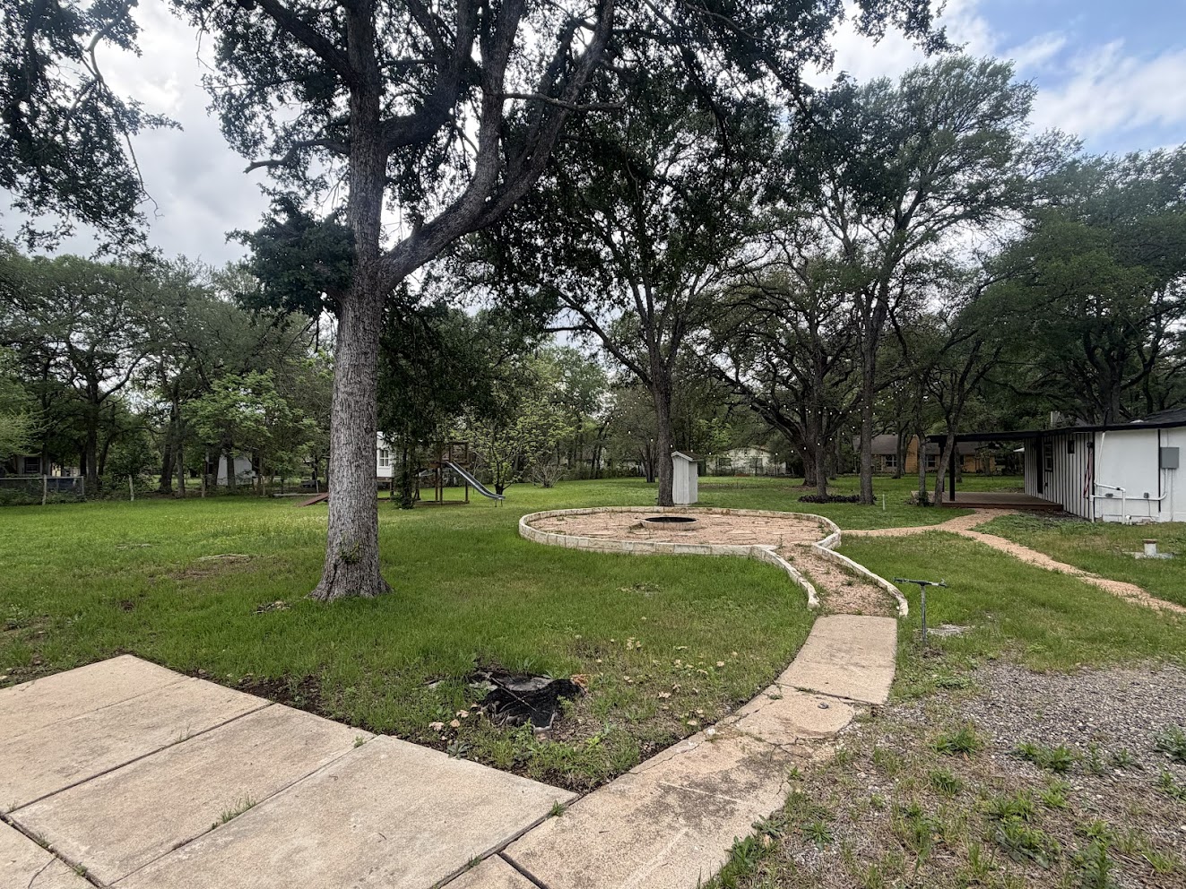 8904 Chisholm Lane Austin, TX 78748 - Photo 5 of 40 Expansive lawn area featuring numerous mature trees, a defined fire pit with a stone border, and a concrete pathway leading to a covered porch structure