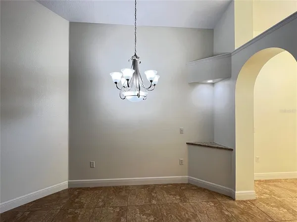 an empty room with chandelier fan and kitchen view