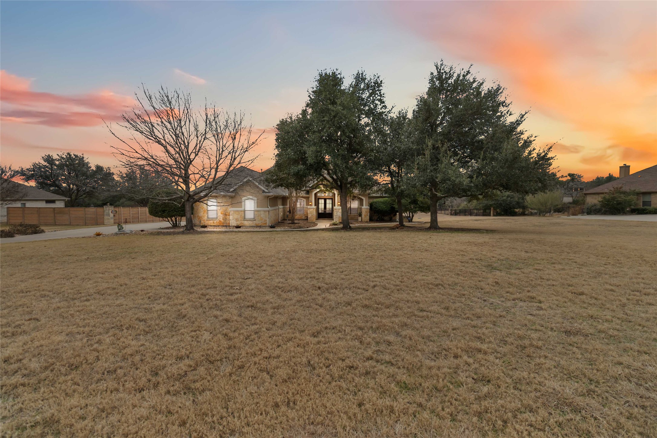A wide front view showcases the home set back on a spacious lot, highlighting the stone exterior, mature trees, and inviting landscaping, all framed by a warm Texas sunset.