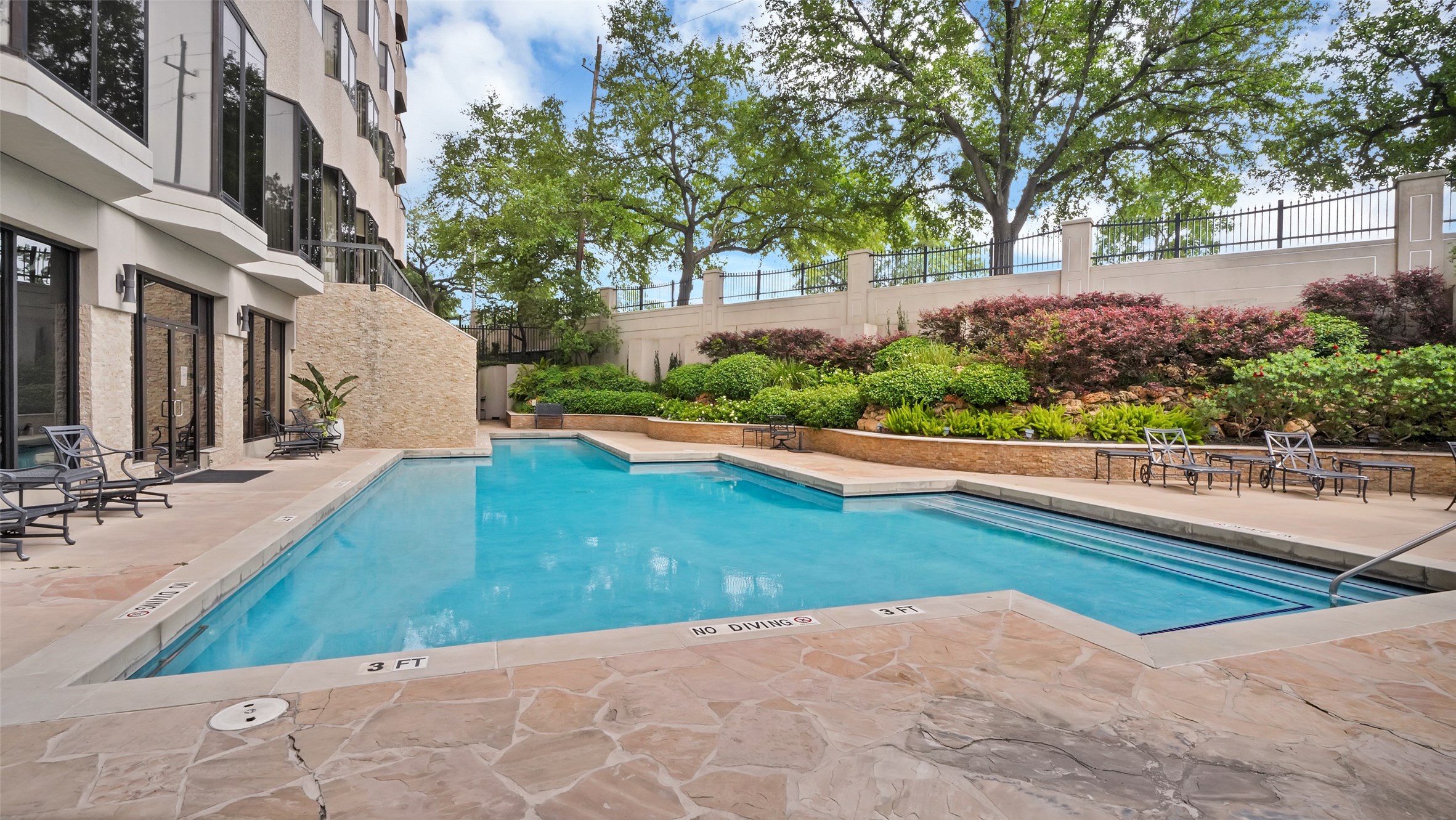 2001 Holcombe Boulevard, Unit 404 Houston, TX 77030 - Photo 18 of 33 This photo showcases a well-maintained outdoor pool area adjacent to a modern building. The pool is surrounded by a stone deck with seating, lush landscaping, and mature trees, creating a private and inviting atmosphere.