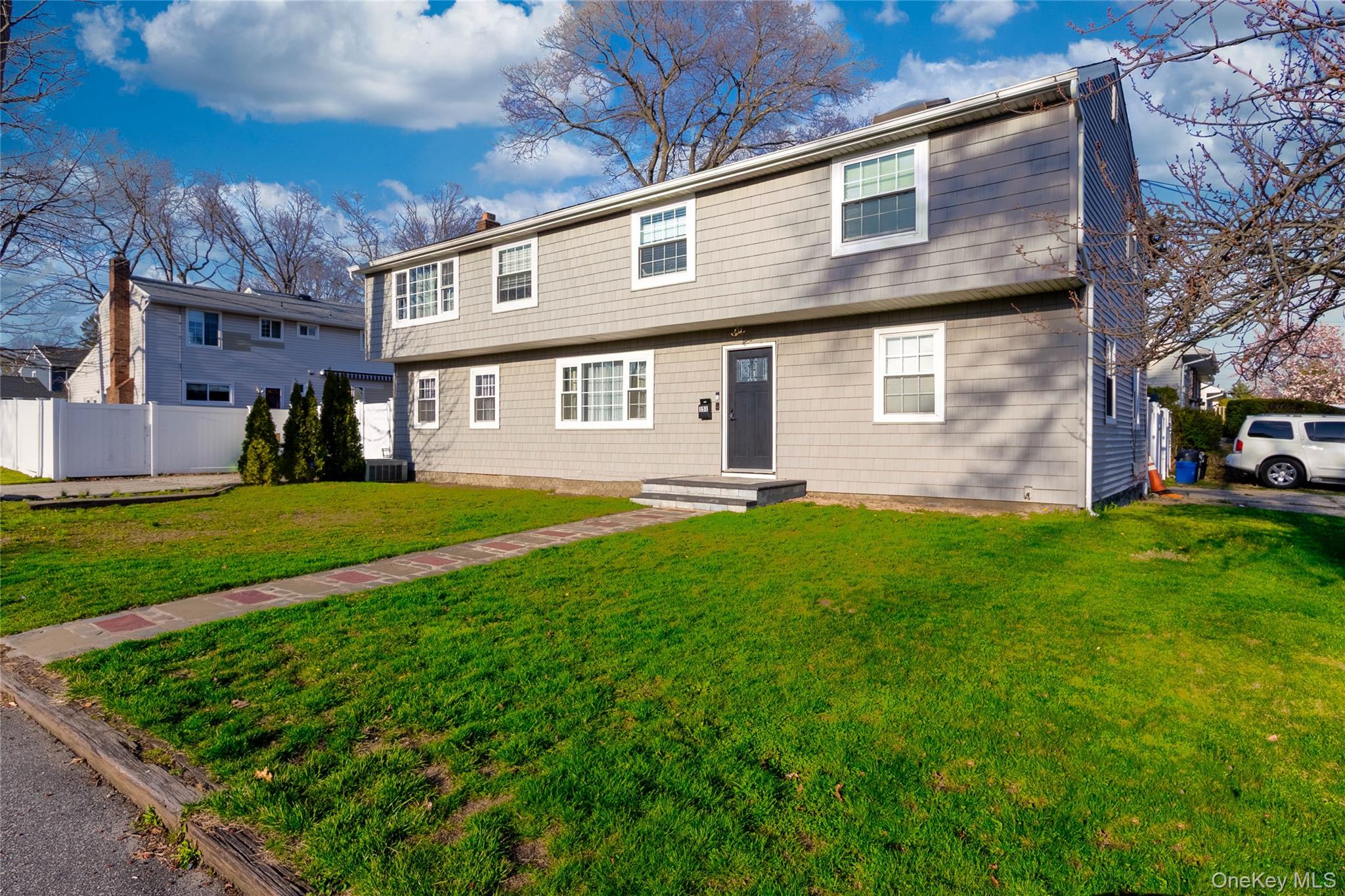151 Cheltenham Road West Babylon, NY 11704 - Photo 2 of 42 The property features a well-maintained lawn, a paved walkway leading to the front entrance, and a light gray exterior with white trim windows