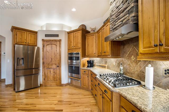 11628 Shaugnessy Road Colorado Springs, CO 80908 - Photo 13 of 50 Kitchen with light stone counters, tasteful backsplash, wall chimney range hood, stainless steel appliances, and light hardwood / wood-style flooring