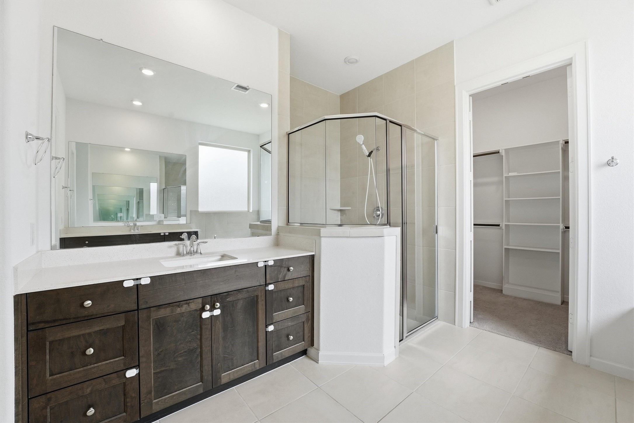 6071 Windward Falls Way Porter, TX 77365 - Photo 19 of 33 This bathroom features a spacious vanity with dark wood cabinets, a large mirror, a glass-enclosed shower, and an adjacent walk-in closet with shelving. The neutral tones and modern fixtures offer a clean, contemporary look.