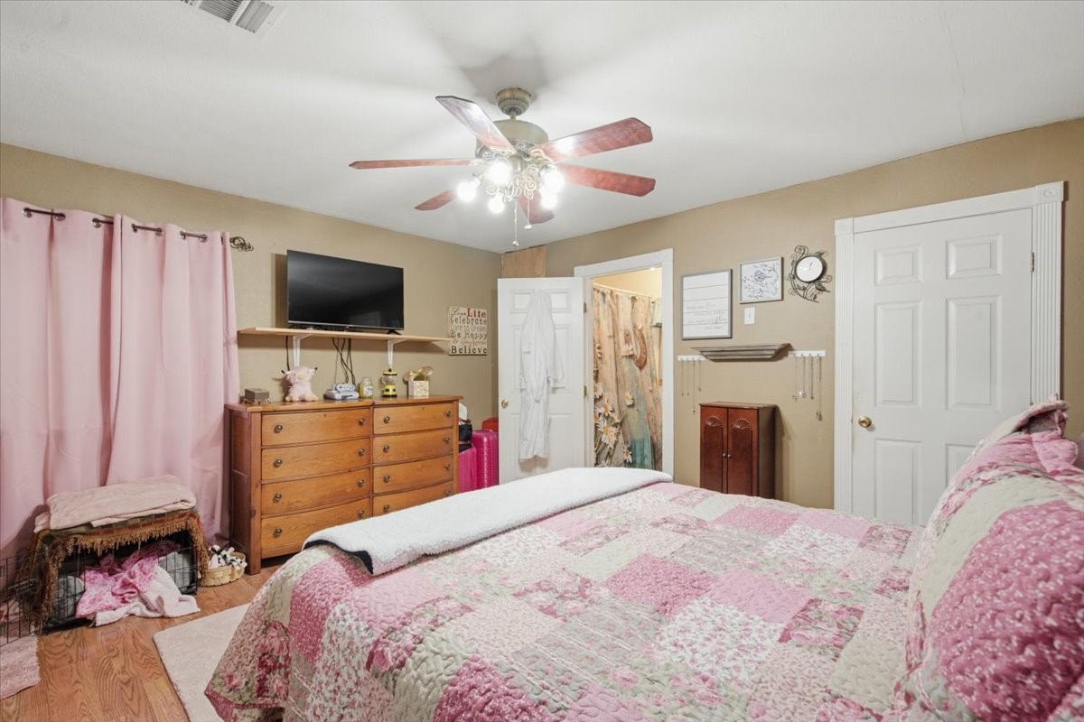 765 Norcross Road Wallis, TX 77485 - Photo 26 of 42 Cozy bedroom with warm tones featuring a ceiling fan, ample lighting, and wood floors. Includes closet space. The room is accented with pink curtains and a patchwork bedspread, adding a homey touch.
