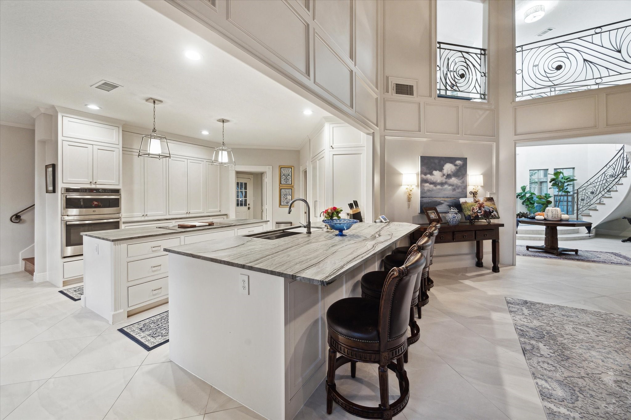 15406 Old Stone Trail Houston, TX 77079 - Photo 42 of 42 This is a spacious, modern kitchen with white cabinetry and high-end appliances, featuring a large marble-topped island with bar seating. The open floor plan offers a view of an elegant staircase and a formal sitting area, all under a two-story ceiling with sophisticated panel detailing.