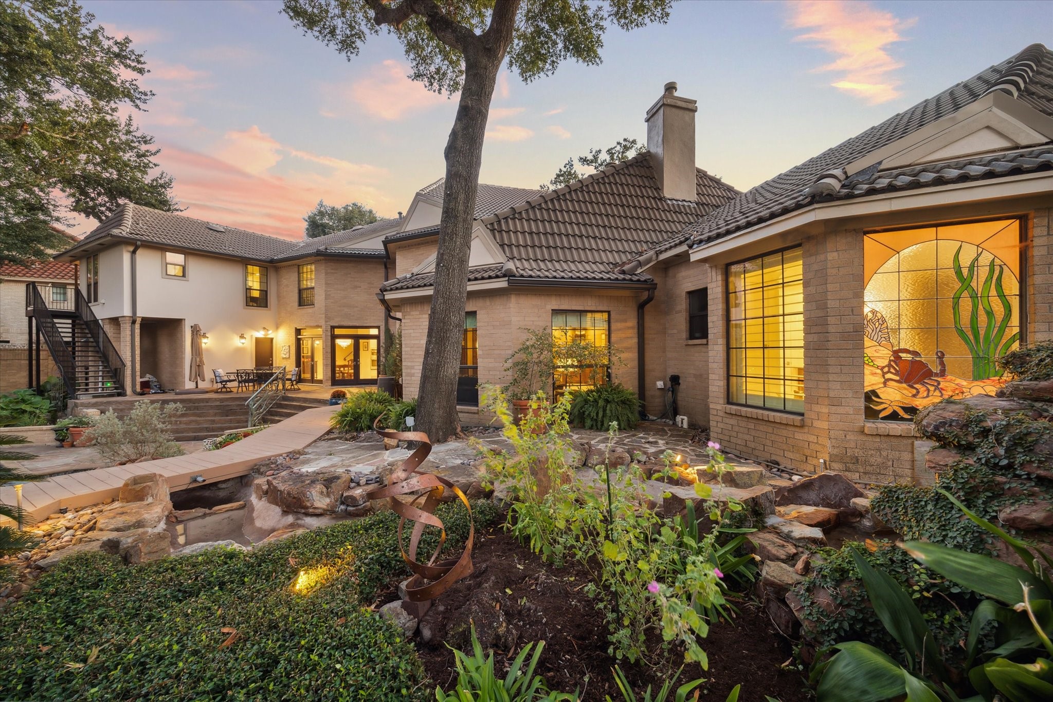 15406 Old Stone Trail Houston, TX 77079 - Photo 35 of 42 This is an elegant two-story home featuring a beautifully landscaped garden with a pathway and outdoor lighting. The residence boasts large windows, a unique stained glass piece, and an inviting outdoor seating area under a picturesque sunset sky.