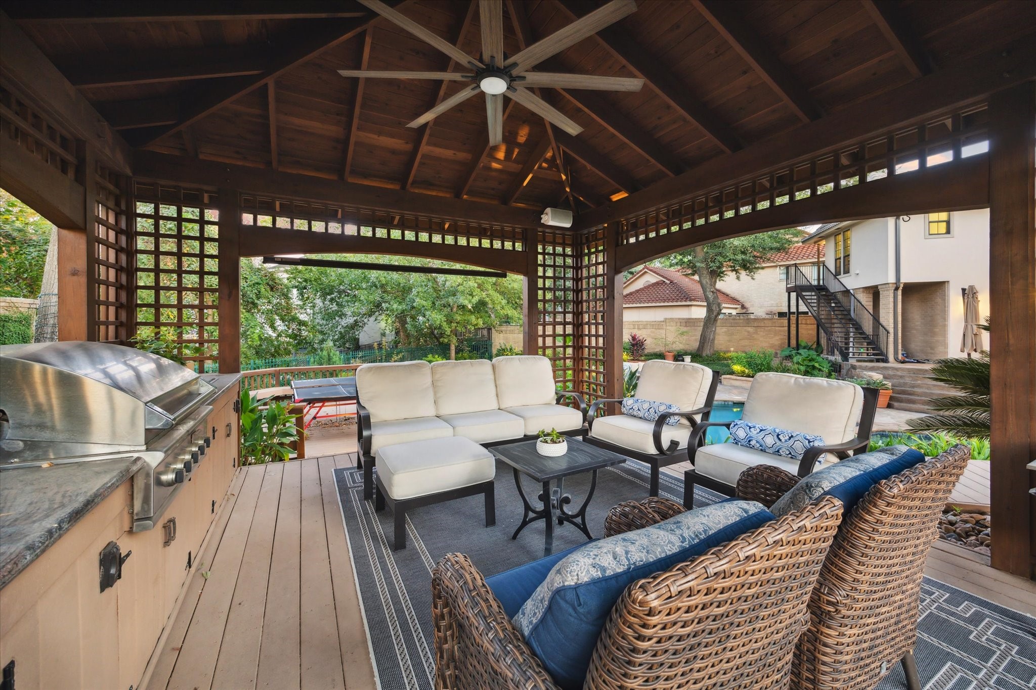 15406 Old Stone Trail Houston, TX 77079 - Photo 36 of 42 This is a cozy, covered outdoor living space with a built-in grill, comfortable seating, and a view of a pool and landscaped garden, perfect for relaxation and entertaining.