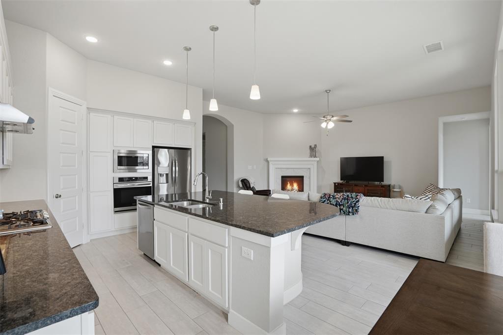 1429 Eclipse Road Aubrey, TX 76227 - Photo 10 of 37 Open concept living space featuring a modern kitchen with a large island, stainless steel appliances, and light-colored flooring, opening into a living room with a fireplace and a ceiling fan