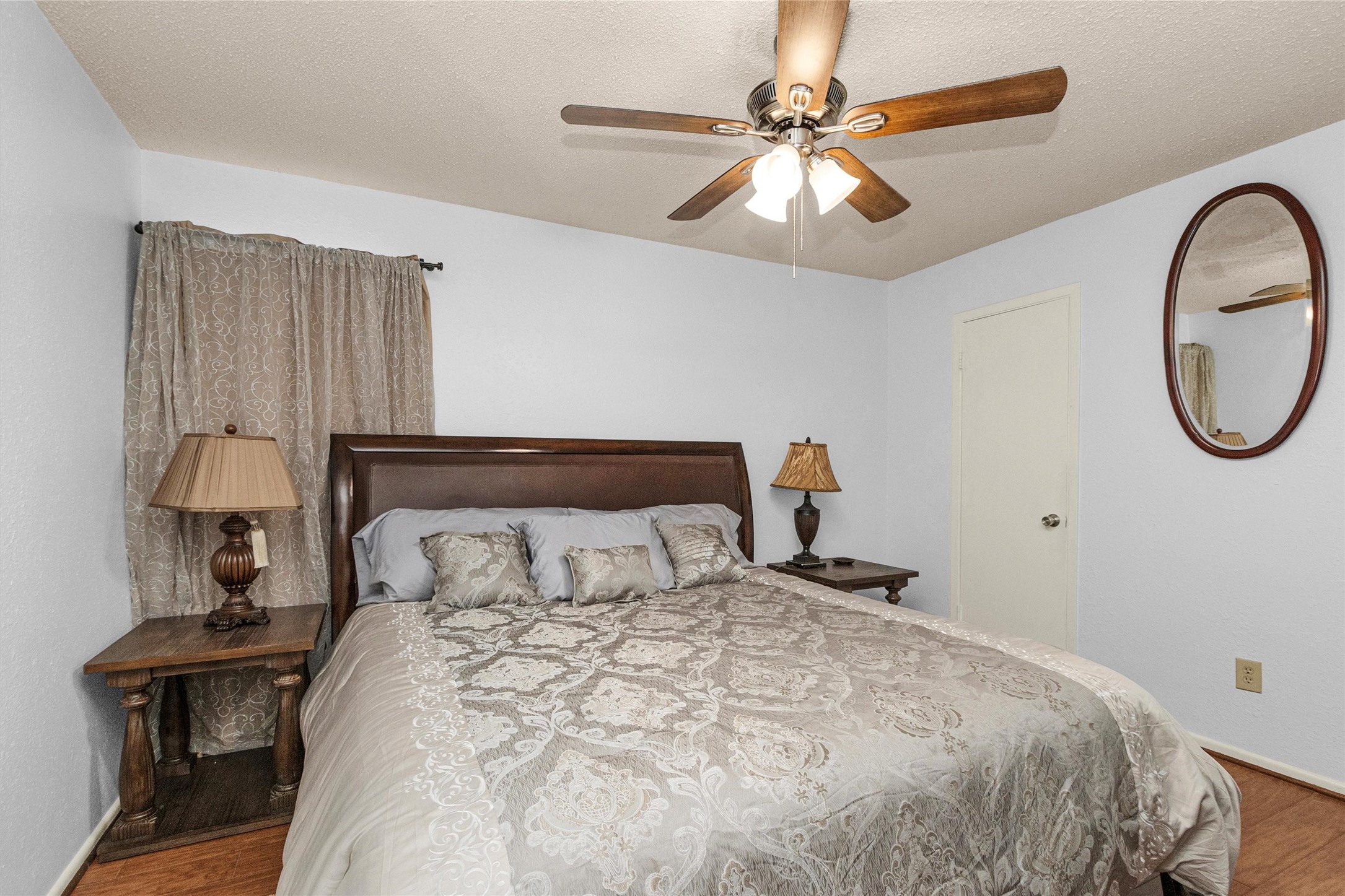 12900 Walden Road, Unit D 411 Montgomery, TX 77356 - Photo 14 of 18 This cozy bedroom invites relaxation with its large bed adorned with a patterned comforter, complemented by matching bedside tables and lamps, a ceiling fan, and a wall mirror, all in warm neutral tones.