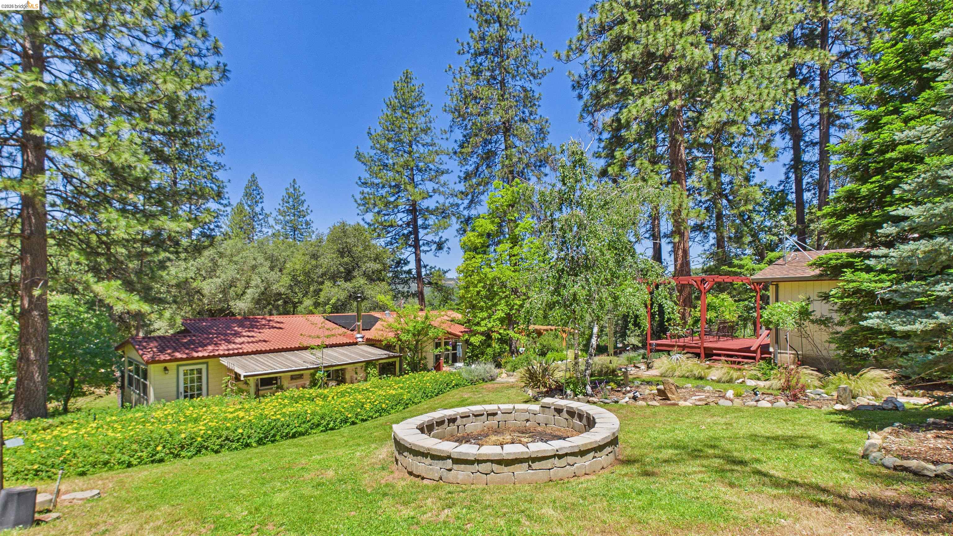 18102 Mt Eaton Road Tuolumne, CA 95379 - Photo 26 of 44 Expansive backyard featuring a stone-clad fire pit, a red wood deck with pergola, and lush green landscaping