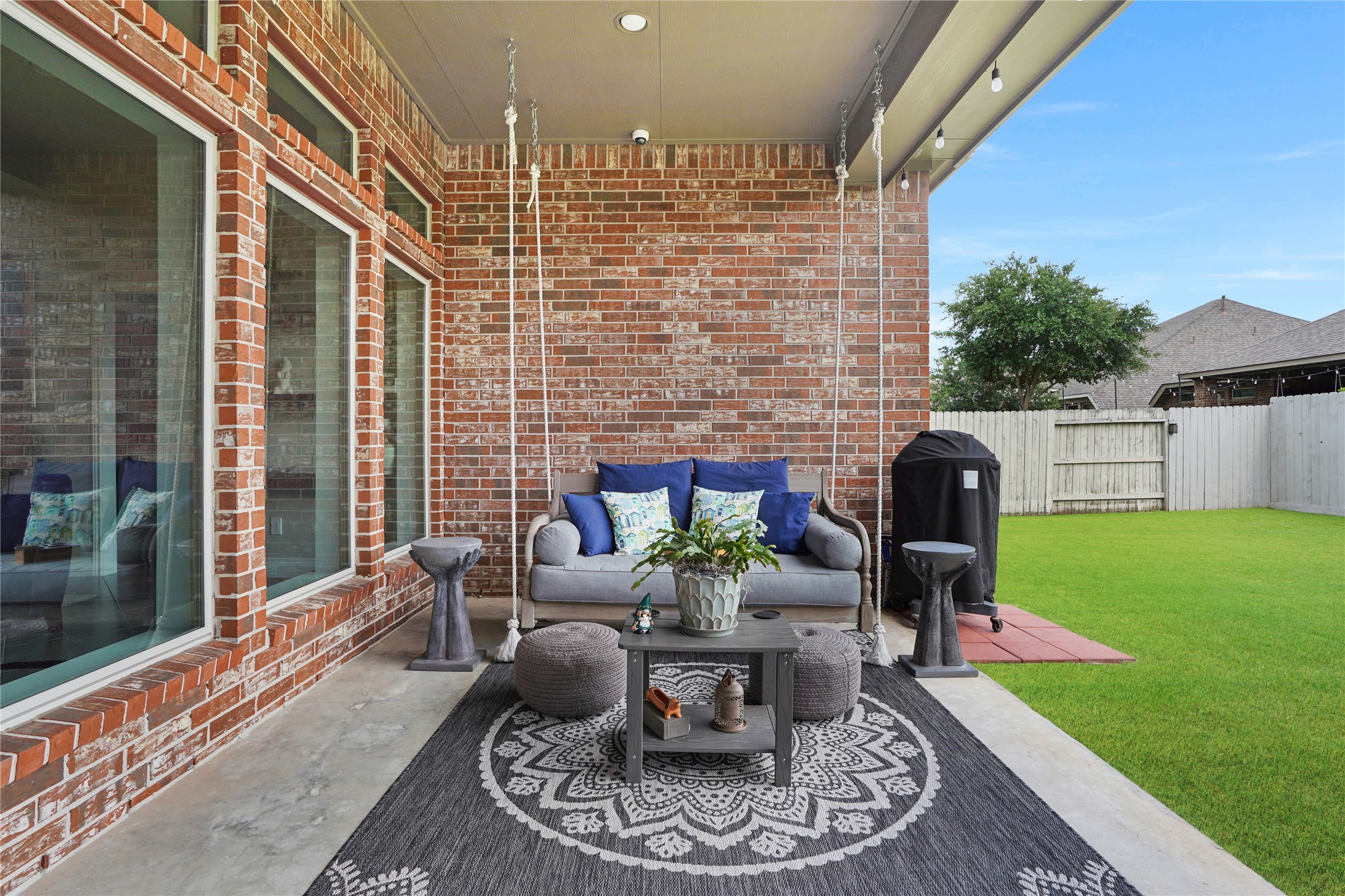 2345 Redwood Ridge Trail Manvel, TX 77578 - Photo 5 of 39 This inviting outdoor patio features a cozy seating area with a swing, surrounded by a brick wall and large windows. It includes a decorative rug, modern side tables, and a grill, all overlooking a well-maintained, fenced backyard. Perfect for relaxing or entertaining.