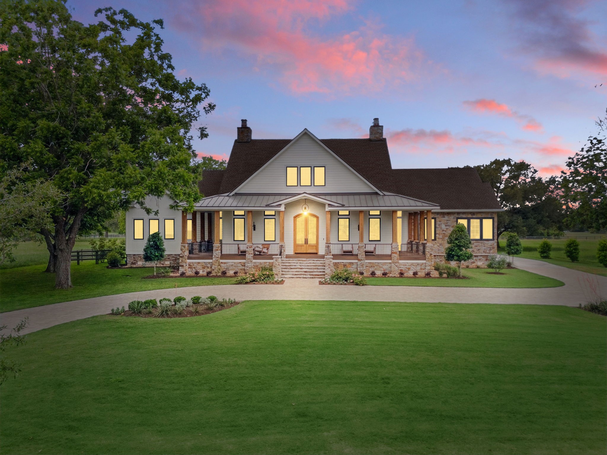 This stunning home features a classic farmhouse design with a spacious front porch, beautiful stone accents, and large windows. It's set against a picturesque backdrop with a well-maintained lawn and mature trees, offering a serene and inviting atmosphere.