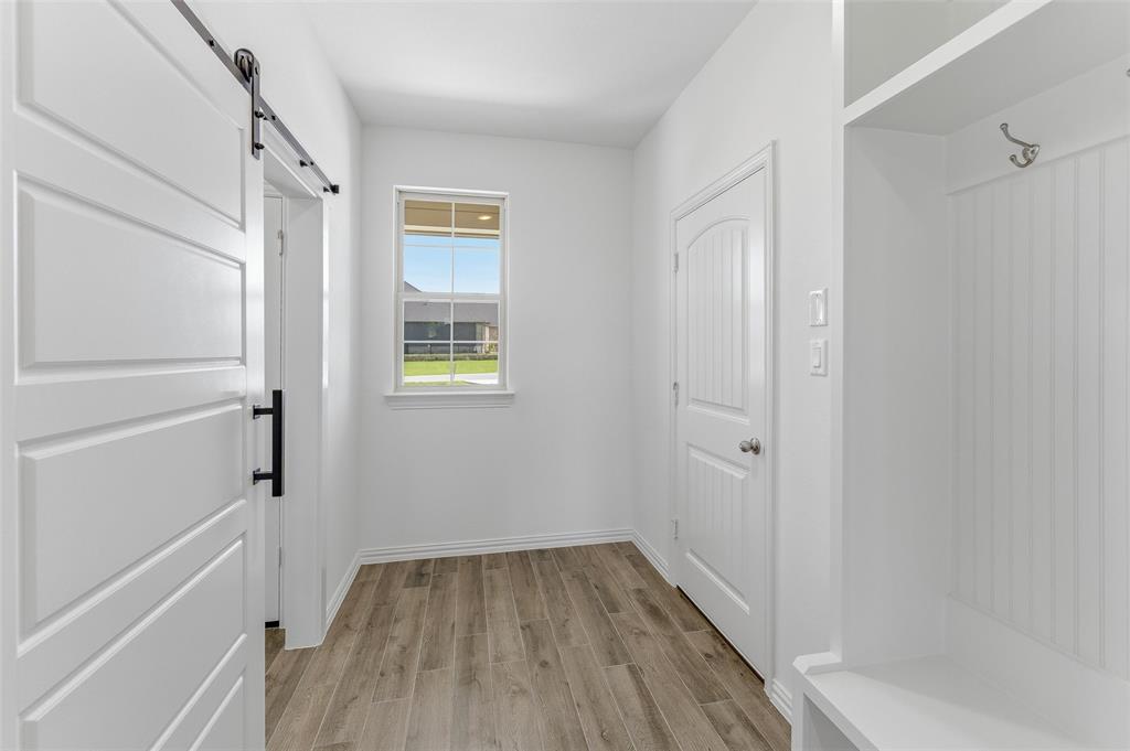 130 Fossil Canyon Drive Rhome, TX 76078 - Photo 11 of 25 Entryway featuring wood-finish flooring, a white paneled barn door with black hardware, a built-in storage bench with beadboard backing and coat hooks, a window with exterior views, and white trim