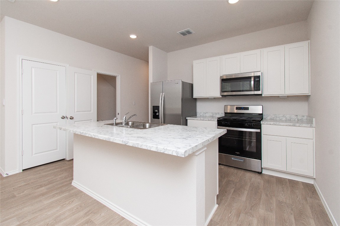 201 Varner Way Lockhart, TX 78644 - Photo 3 of 16 Kitchen featuring appliances with stainless steel finishes, light countertops, a kitchen island with sink, white cabinets, and light wood-style flooring