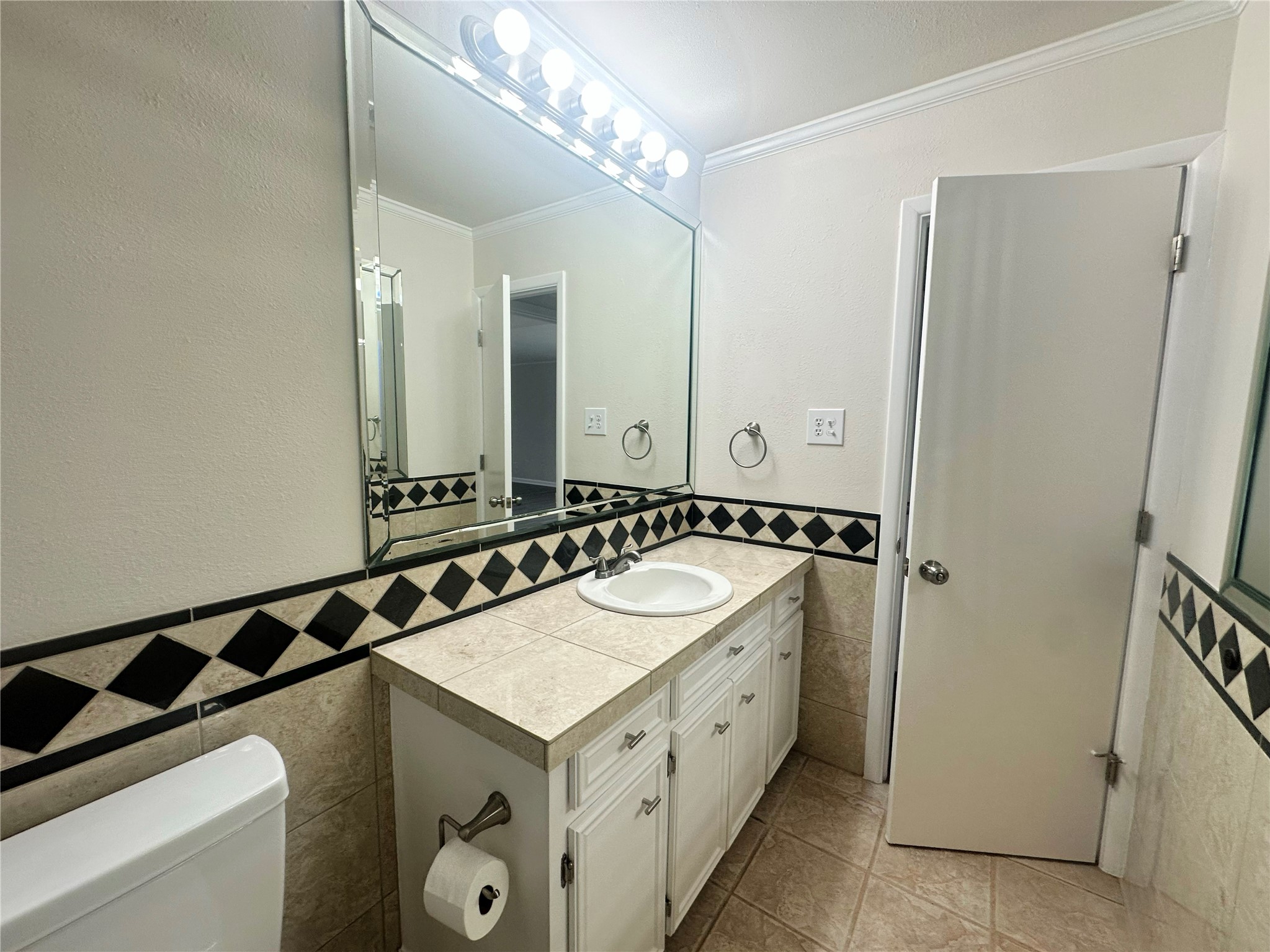 130 Bay Ridge Drive Baytown, TX 77523 - Photo 11 of 20 This bathroom features a large mirror with bright vanity lighting, a tiled countertop with a single sink, and decorative black and white tile accents. There's ample cabinet storage and a modern, clean design.