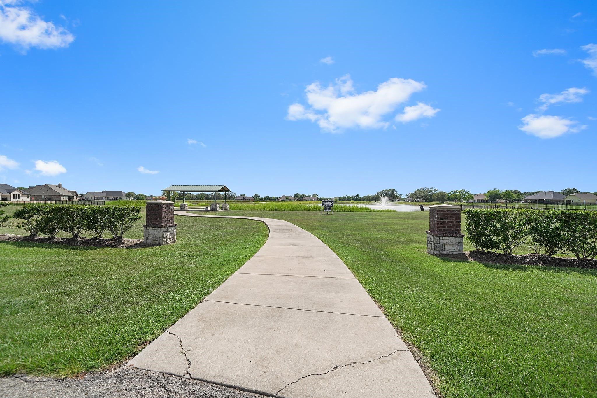351 Lily Lane Rosharon, TX 77583 - Photo 3 of 5 This photo showcases a scenic community area with a paved walkway leading to a gazebo and a pond with a fountain. Neatly trimmed hedges and spacious green lawns create a welcoming outdoor space, ideal for relaxation and leisure activities.