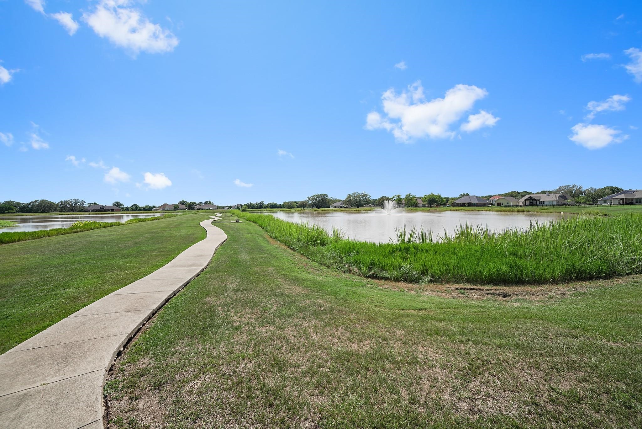 351 Lily Lane Rosharon, TX 77583 - Photo 4 of 5 This photo showcases a serene pathway alongside a pond with a fountain, surrounded by lush greenery and distant houses, under a bright blue sky. Ideal for tranquil walks and enjoying nature.