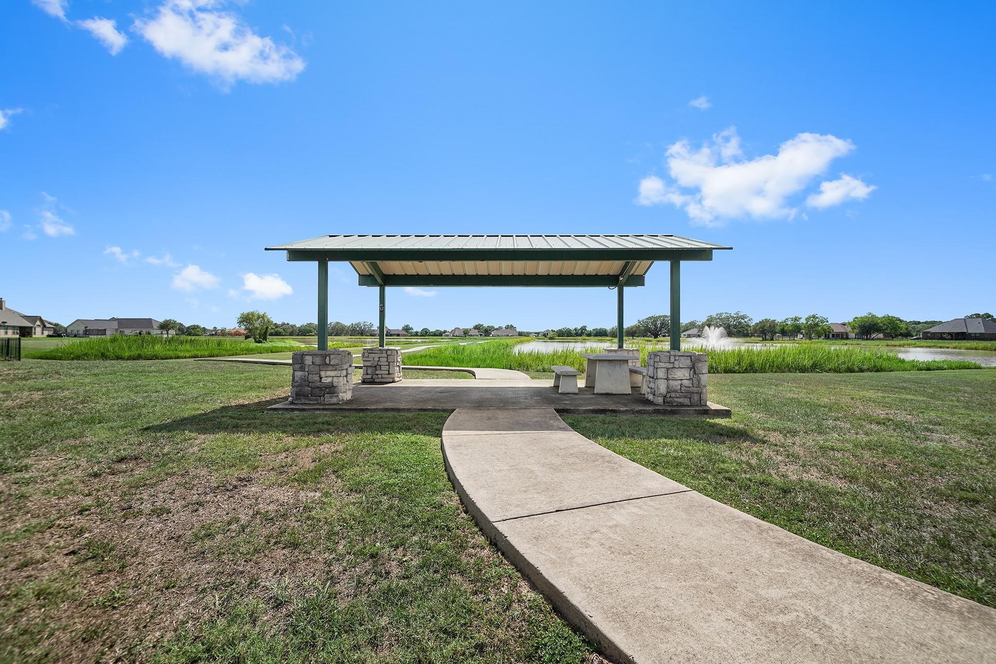 351 Lily Lane Rosharon, TX 77583 - Photo 5 of 5 A charming outdoor pavilion with a metal roof and stone pillars is set on a grassy lawn. A curved pathway leads to it, offering seating and views of a serene pond with a fountain, perfect for relaxation or gatherings.