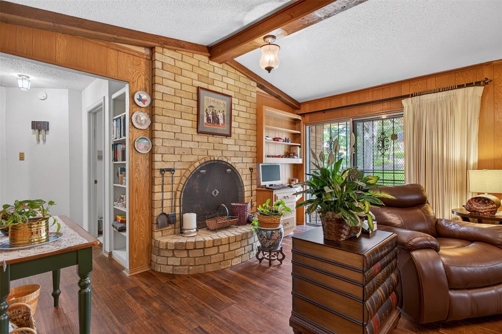 6010 Hunters View Lane Dallas, TX 75232 - Photo 14 of 28 Spacious living area featuring a brick fireplace with a raised hearth, wood-finish flooring, wood paneling, exposed wood beams, and built-in shelving