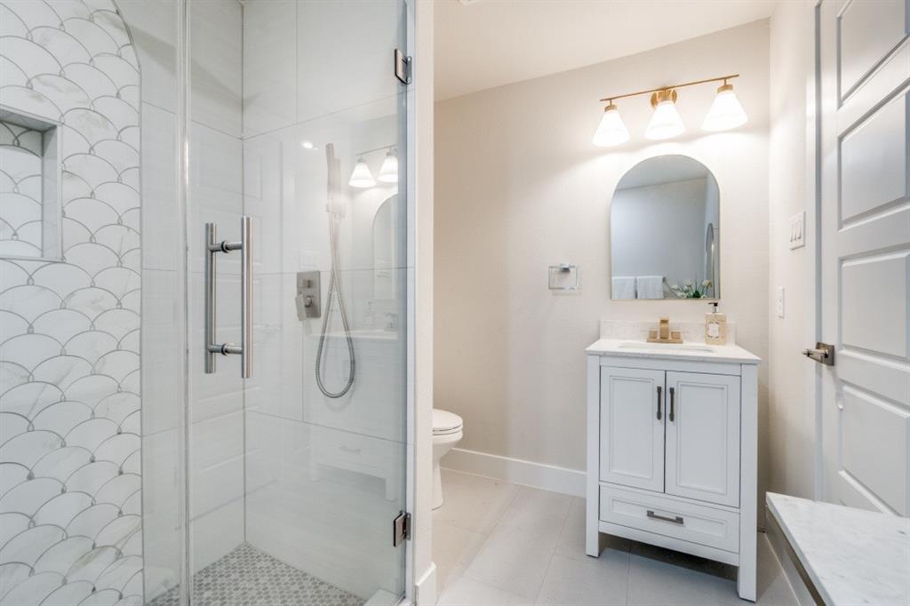 7115 Meadow Road Dallas, TX 75230 - Photo 9 of 22 The bathroom features a glass-enclosed shower with decorative scallop-patterned tile, a white vanity with a rectangular sink, and an arched mirror with a three-light fixture