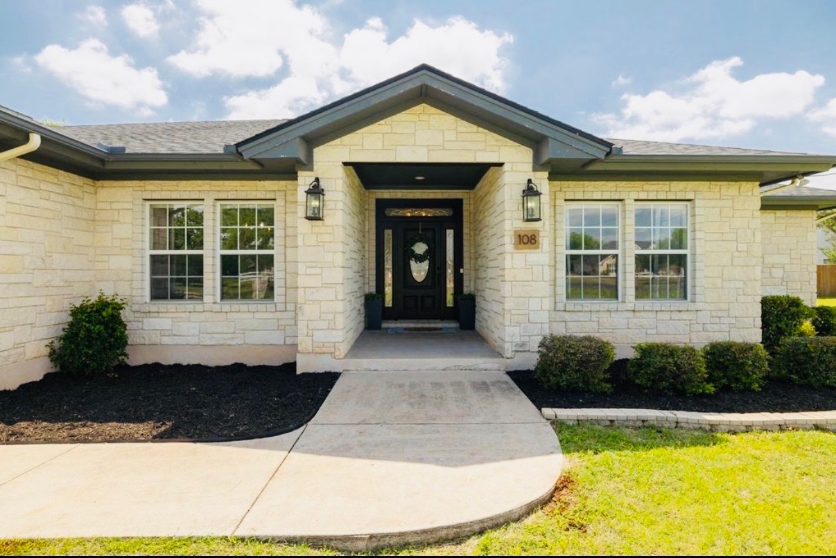 108 Kyle Lane Georgetown, TX 78633 - Photo 23 of 26 Limestone exterior featuring a dark paneled entry door with decorative glass, dual exterior sconces, a dark shingle roof, and a concrete pathway
