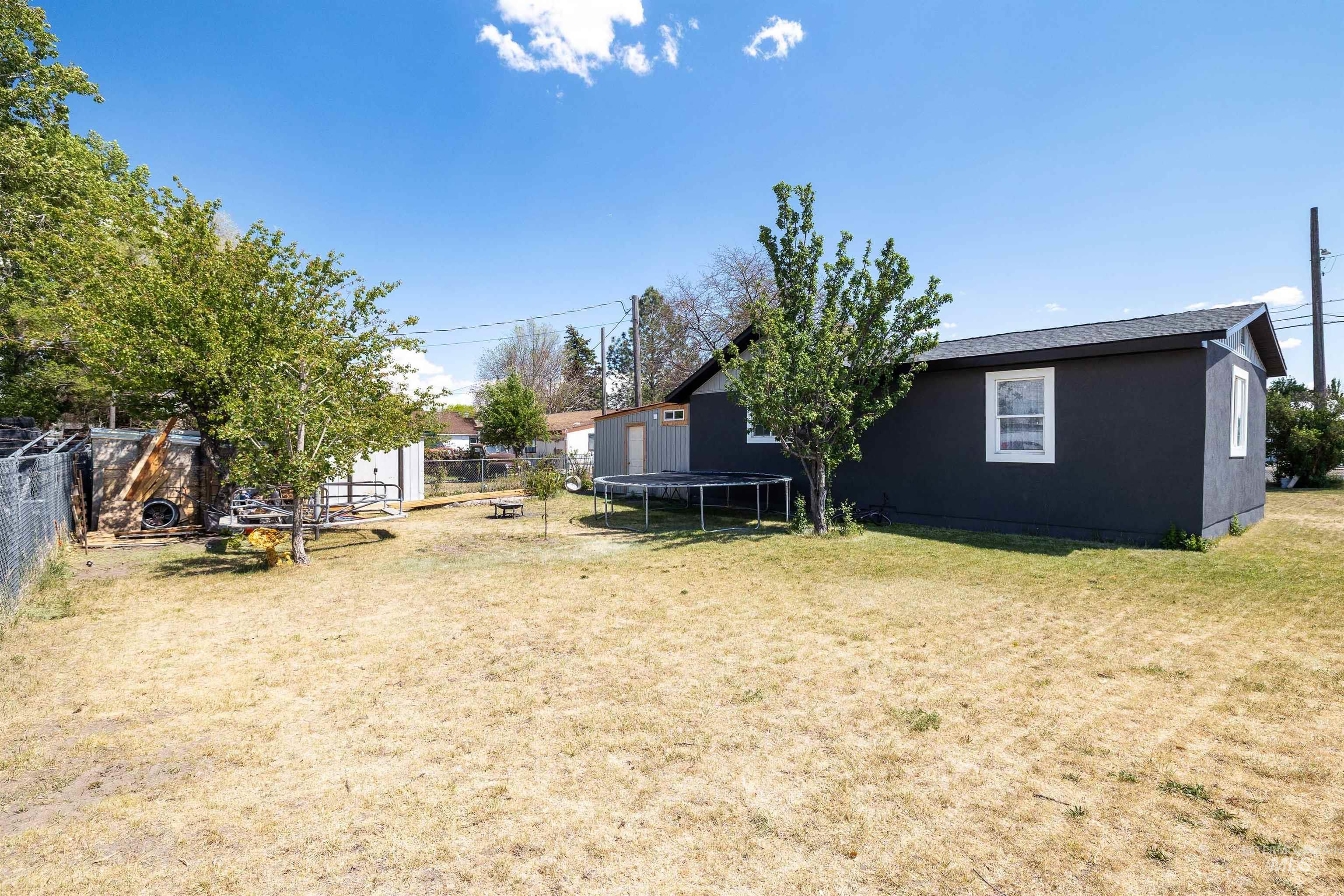 102 East 6th Street Rupert, ID 83350 - Photo 22 of 27 Expansive backyard featuring a dark-colored exterior structure with white trim windows, mature trees, and a chain-link fence