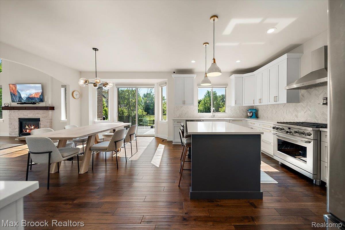 1942 Nottingham Drive Rochester, MI 48307 - Photo 27 of 97 Open-concept living space featuring wood-finish flooring, a kitchen with white cabinetry and a dark-toned island, a herringbone tile backsplash, a stainless steel range with a ventilation hood, and a fireplace with a stone surround