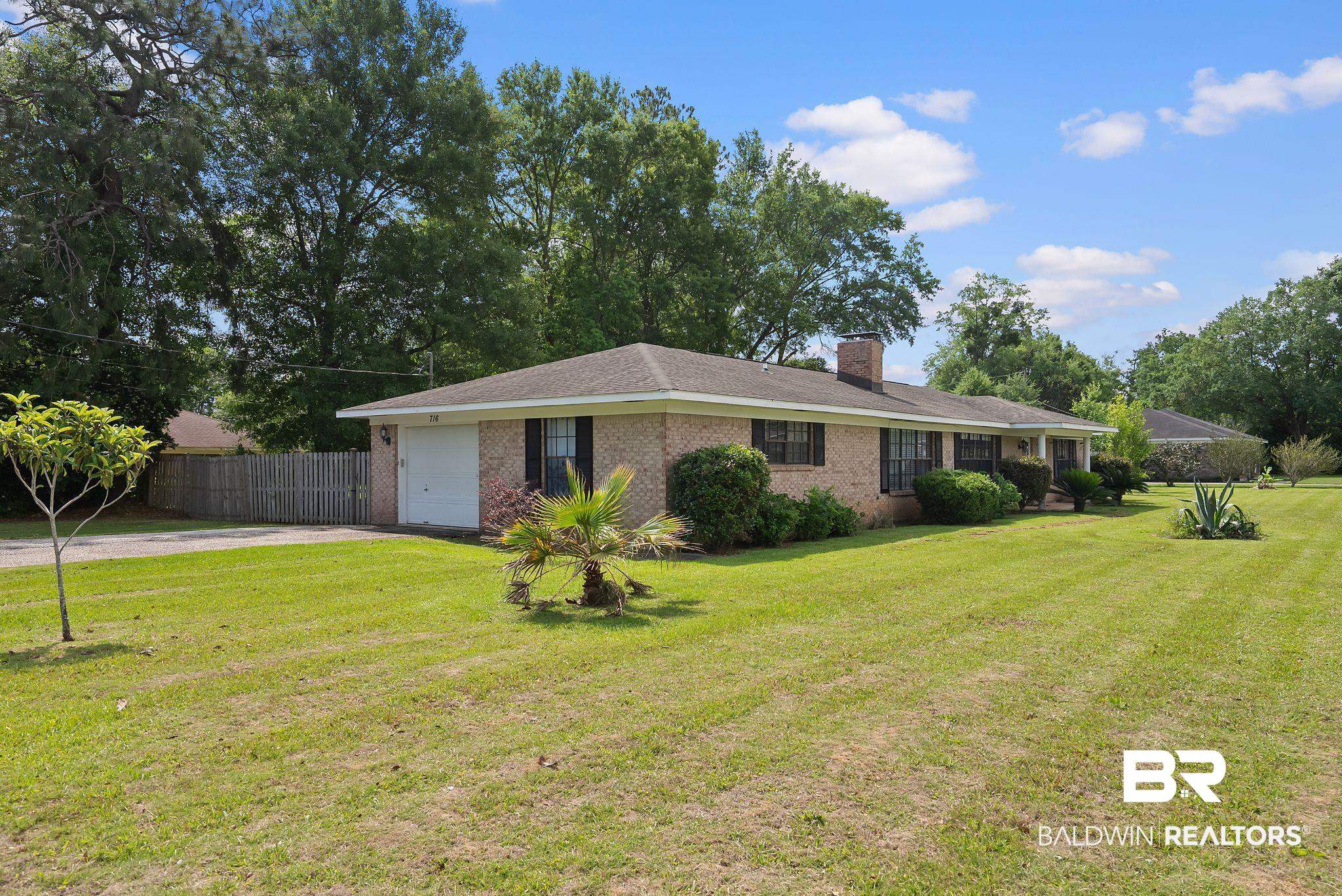 716 North Cedar Street Foley, AL 36535 - Photo 2 of 25 Brick ranch-style residence featuring an attached single-car garage, dark window shutters, established foundation plantings, a prominent brick chimney, and a large lawn with mature trees