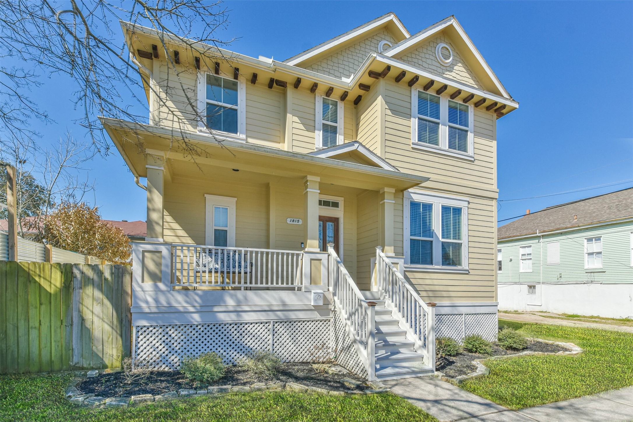 Charming two-story yellow home with a welcoming front porch, white railings, and a manicured lawn. The house features multiple large windows and architectural details under a clear blue sky.