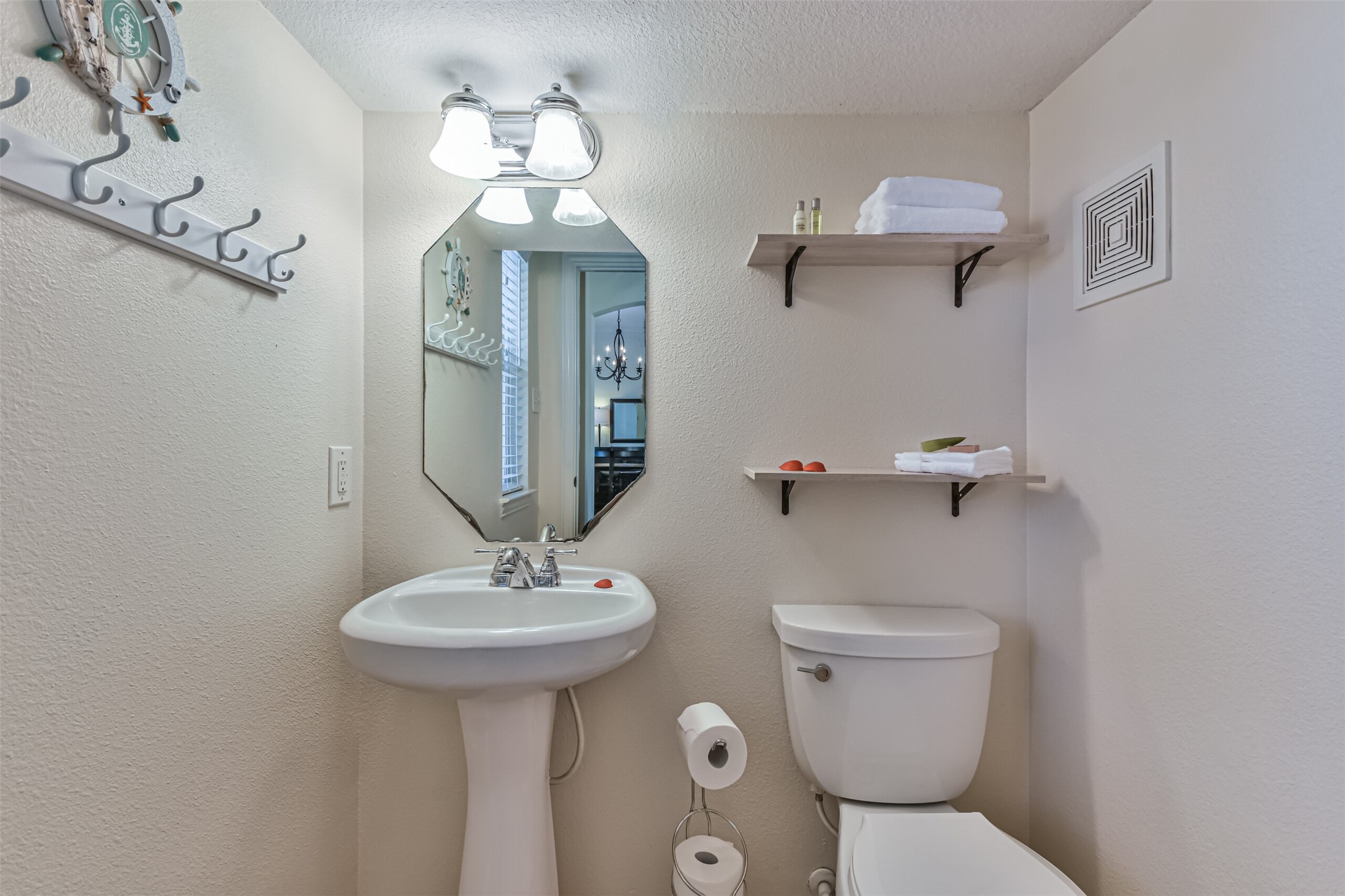 1815 28th Street Galveston, TX 77550 - Photo 11 of 42 This bathroom features a compact design with a pedestal sink, a toilet, and wall-mounted shelves for storage. A decorative mirror and overhead lighting add charm and functionality.