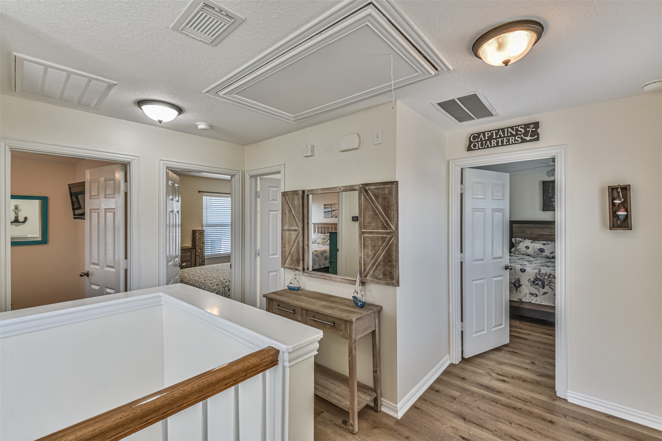 1815 28th Street Galveston, TX 77550 - Photo 12 of 42 This photo shows a bright, welcoming hallway with wooden flooring and light-colored walls. There are three doorways leading to bedrooms. A decorative console table with a mirror and nautical-themed accents adds charm to the space.