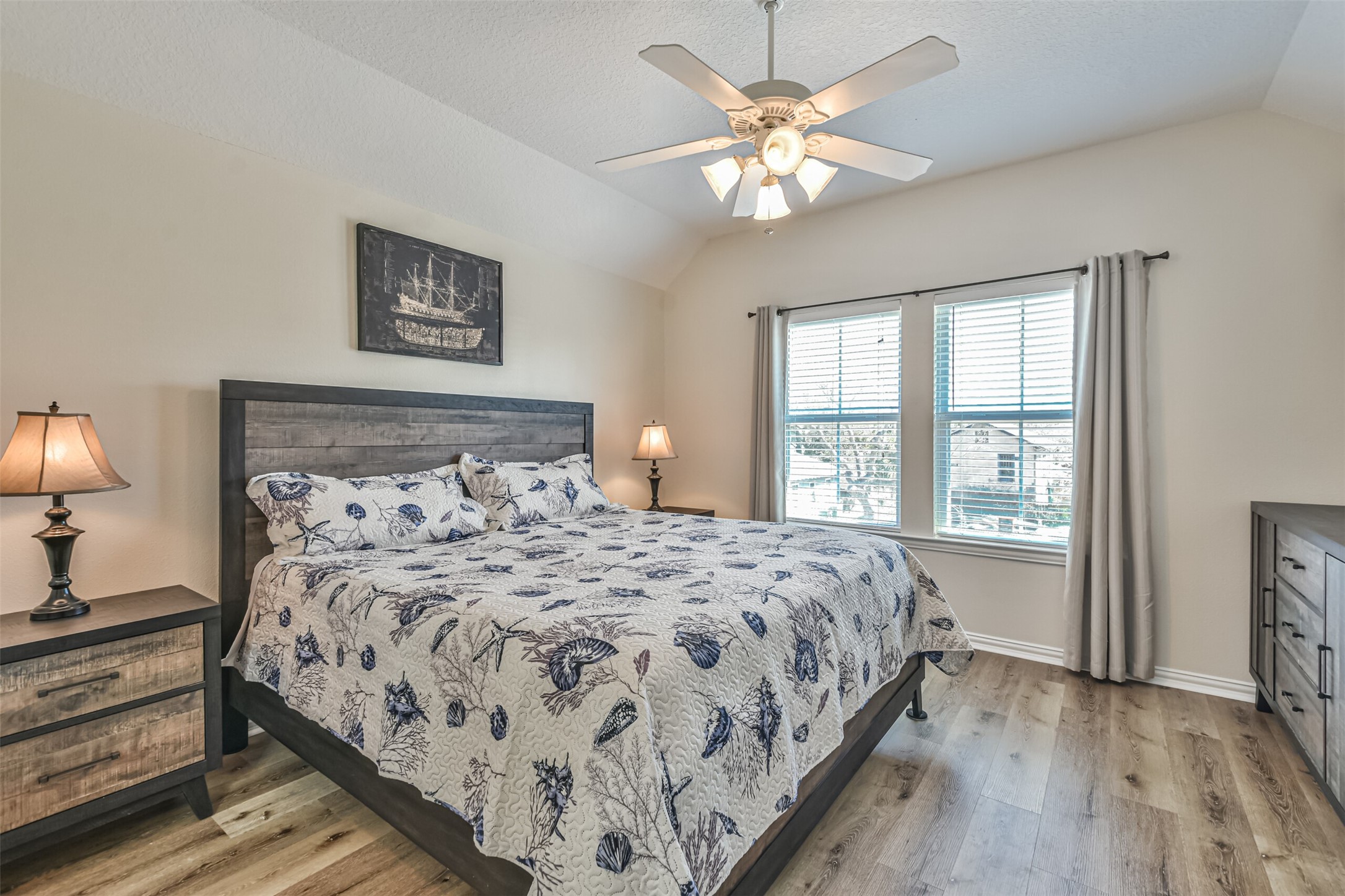 1815 28th Street Galveston, TX 77550 - Photo 13 of 42 This cozy bedroom features a wooden bed frame with a floral bedspread, matching bedside tables with lamps, and a ceiling fan. Large windows provide ample natural light, complemented by neutral walls and wood flooring.
