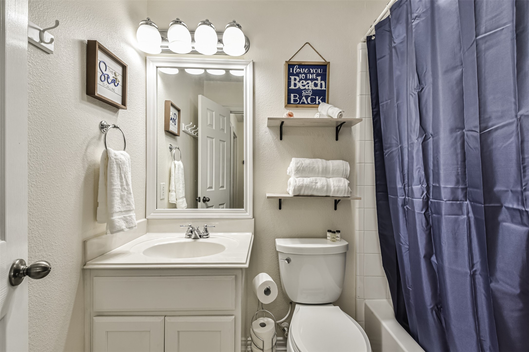 1815 28th Street Galveston, TX 77550 - Photo 18 of 42 This bathroom features a clean, modern design with a white vanity, bright lighting, and a large mirror. It includes a toilet, bathtub with a blue shower curtain, and decorative shelving with towels. The walls have light, neutral colors and charming beach-themed decor.