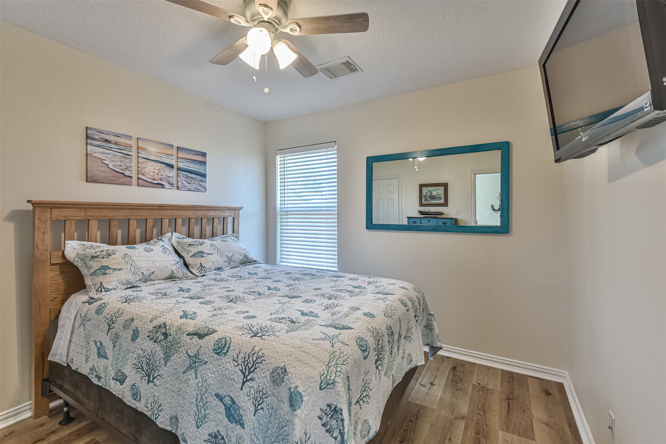 1815 28th Street Galveston, TX 77550 - Photo 19 of 42 Cozy bedroom with a wooden bed frame, ocean-themed bedding, and wall art. Features a ceiling fan, large mirror, and wall-mounted TV. Natural light flows through a window with blinds, complementing the neutral walls and wood flooring.