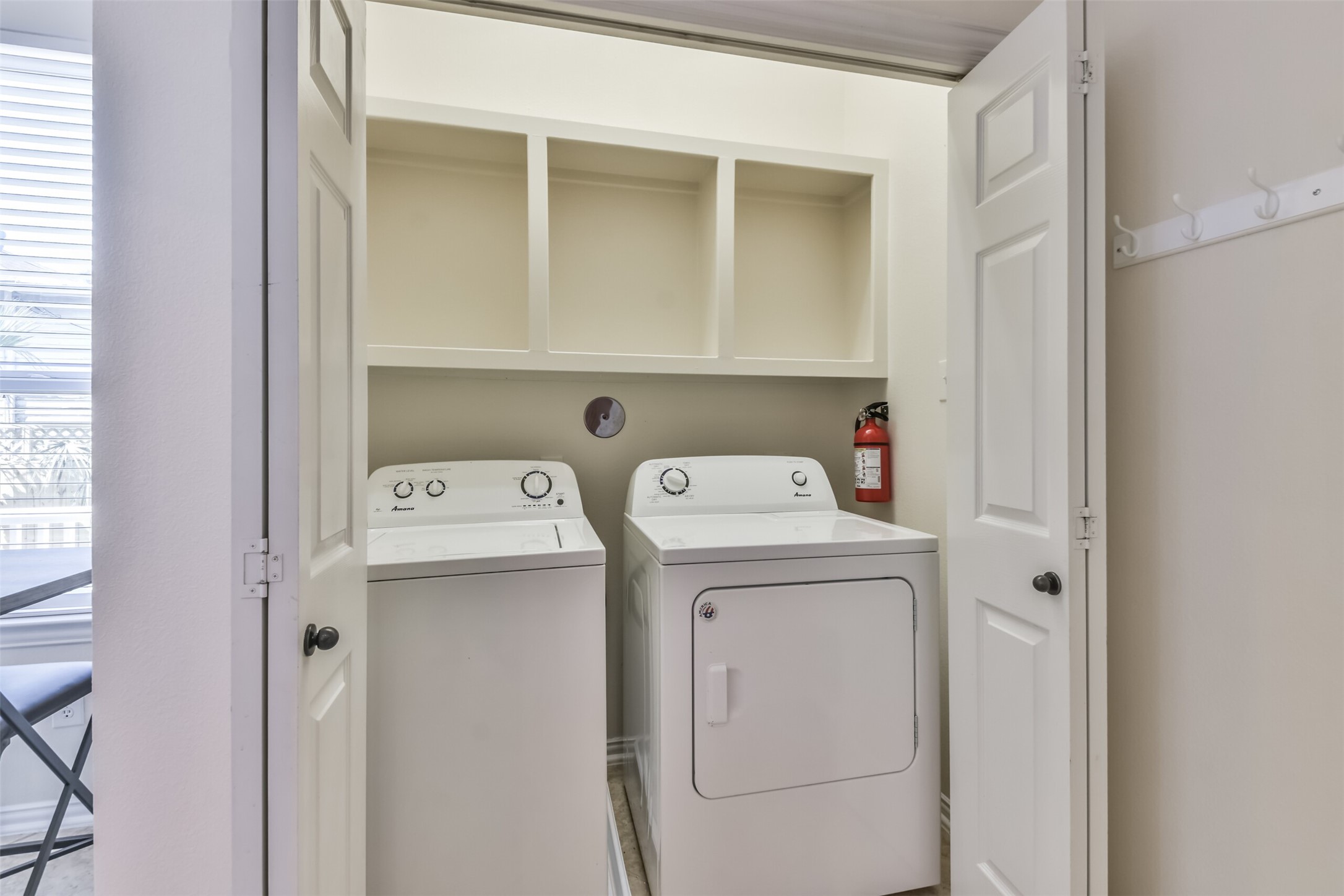1815 28th Street Galveston, TX 77550 - Photo 21 of 42 This photo shows a compact laundry area with a washer and dryer set, open shelving above for storage, and a fire extinguisher nearby. The space is enclosed by folding doors and is well-lit by natural light from a nearby window.