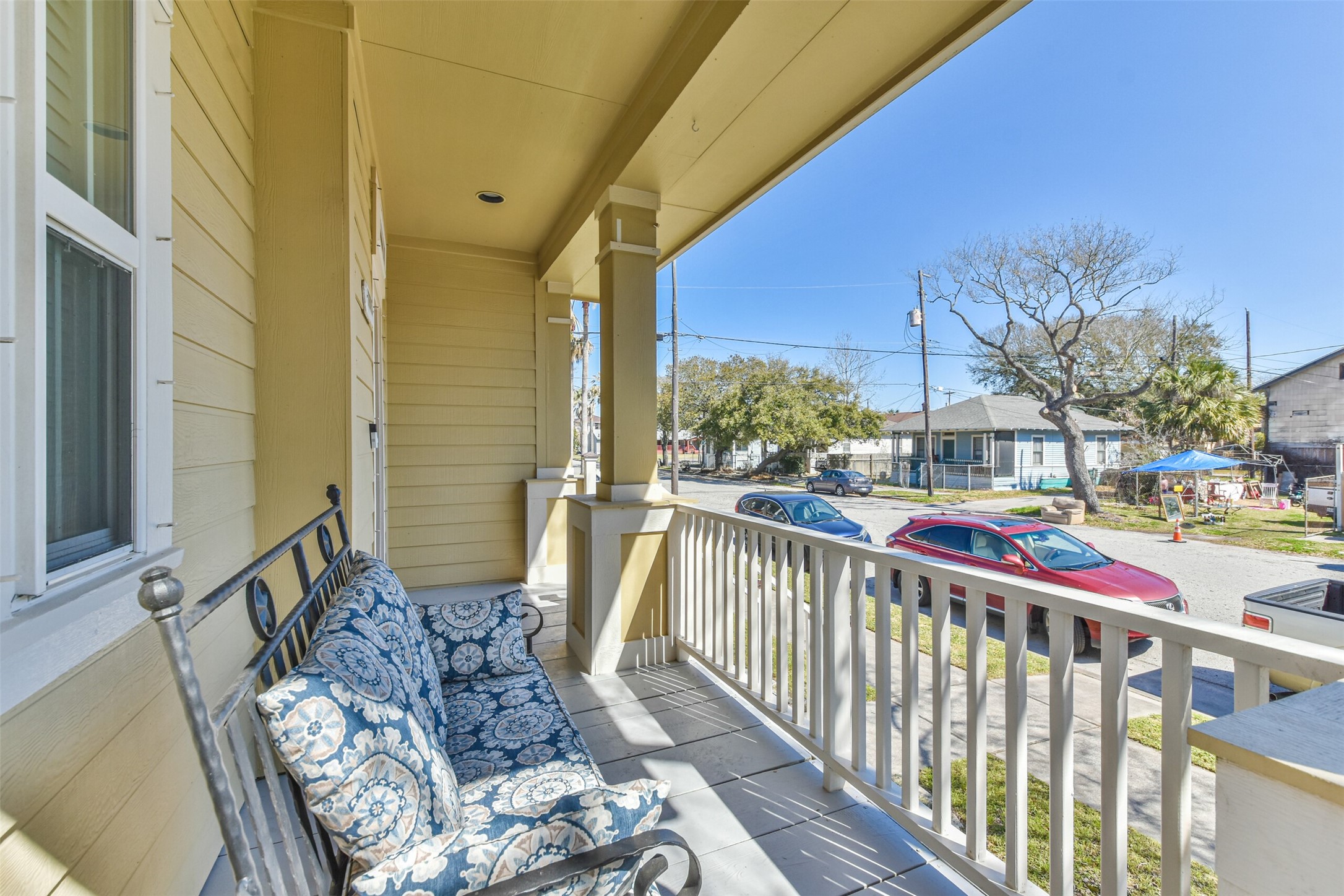 1815 28th Street Galveston, TX 77550 - Photo 25 of 42 This photo showcases a cozy front porch with a comfortable cushioned bench, perfect for relaxing. The porch features classic railing and overlooks a quiet residential street with trees and neighboring houses.